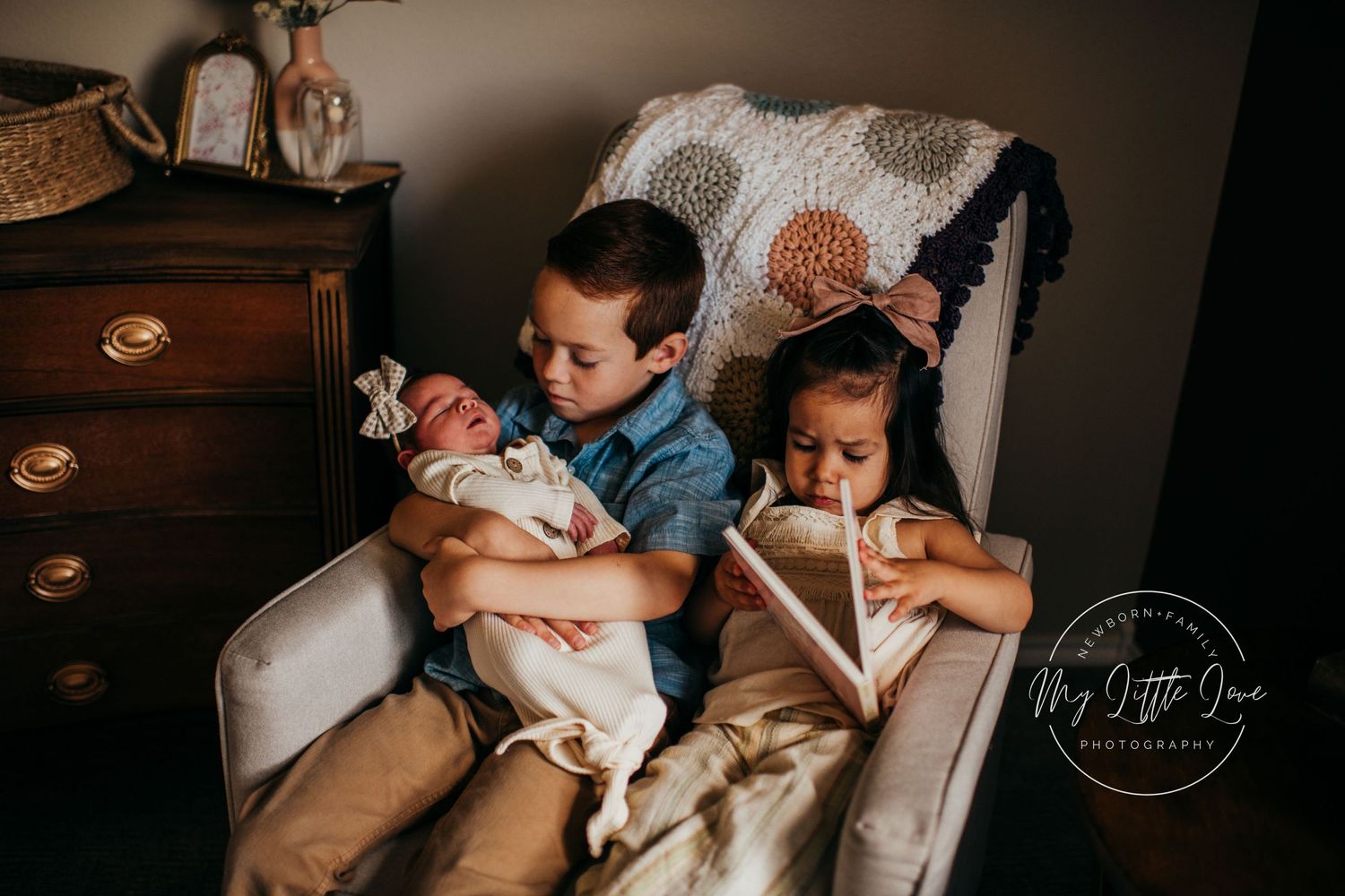 Young siblings sitting in cream armchair while holding a baby.