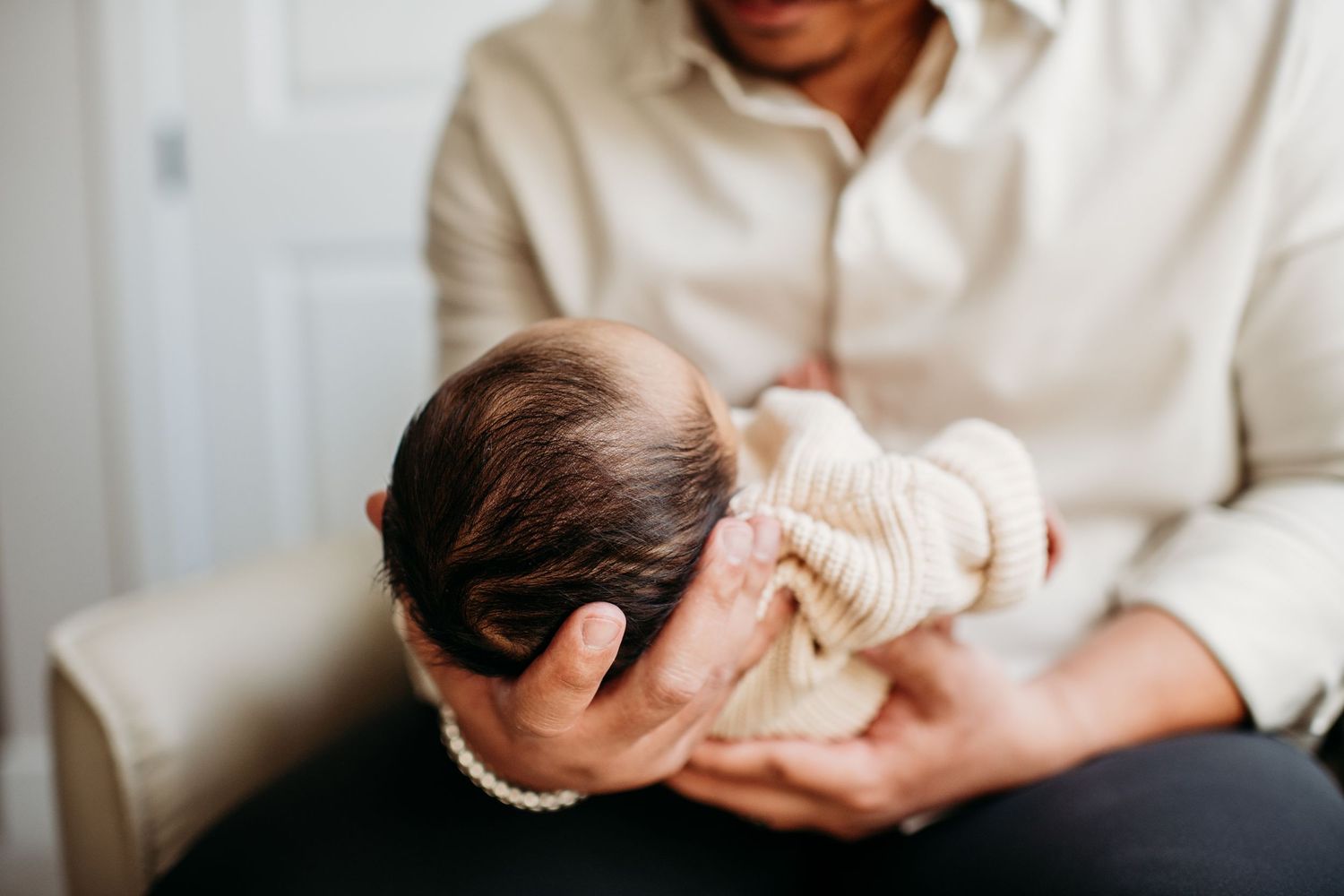 A parent holds a newborn baby swaddled in a cream-colored blanket while sitting on a couch.