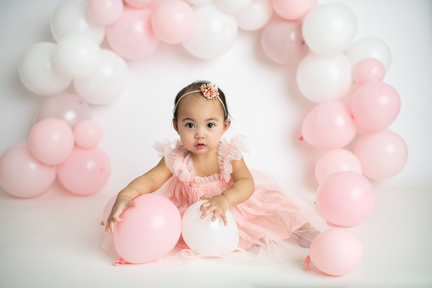 Baby girl in pink ruffle dress sits among pink and white balloons during first birthday cake smash photo session.