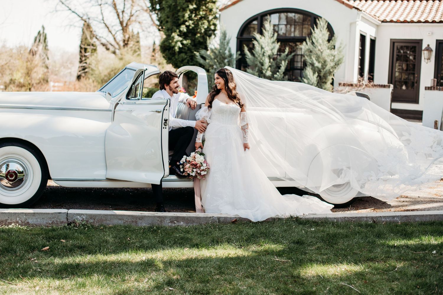 A classic vintage white car parked outside a Mediterranean-style building with flowing white fabric caught in the breeze.