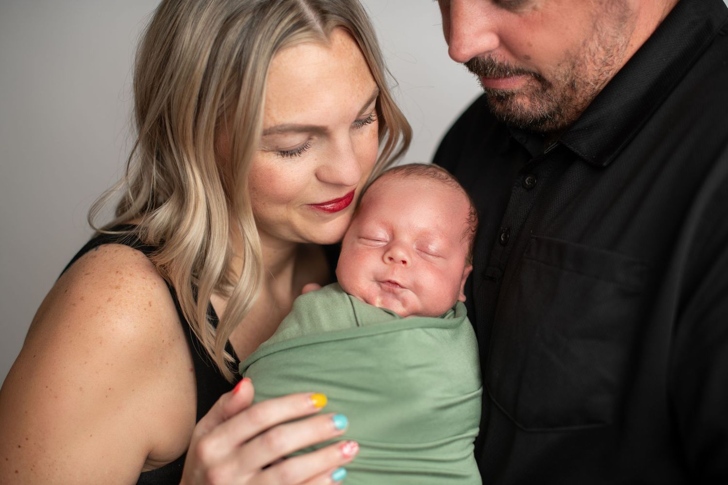A couple tenderly holds their sleeping newborn baby wrapped in a green swaddle blanket in a studio portrait.