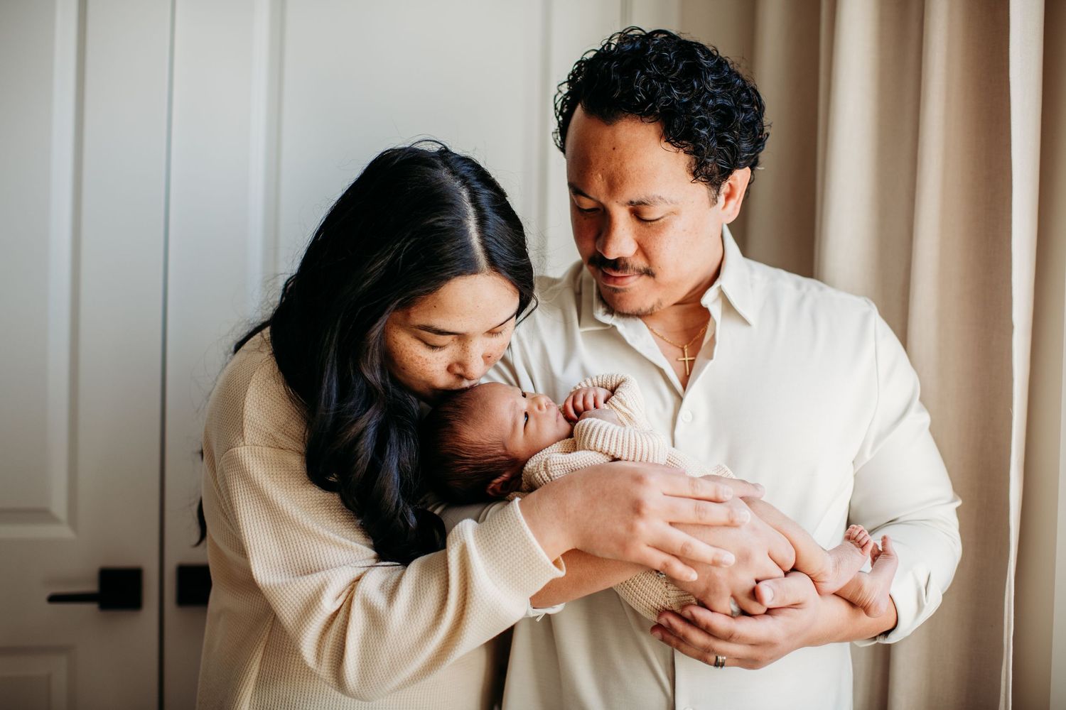 New parents sharing a tender moment with their sleeping newborn baby inside a softly lit room.