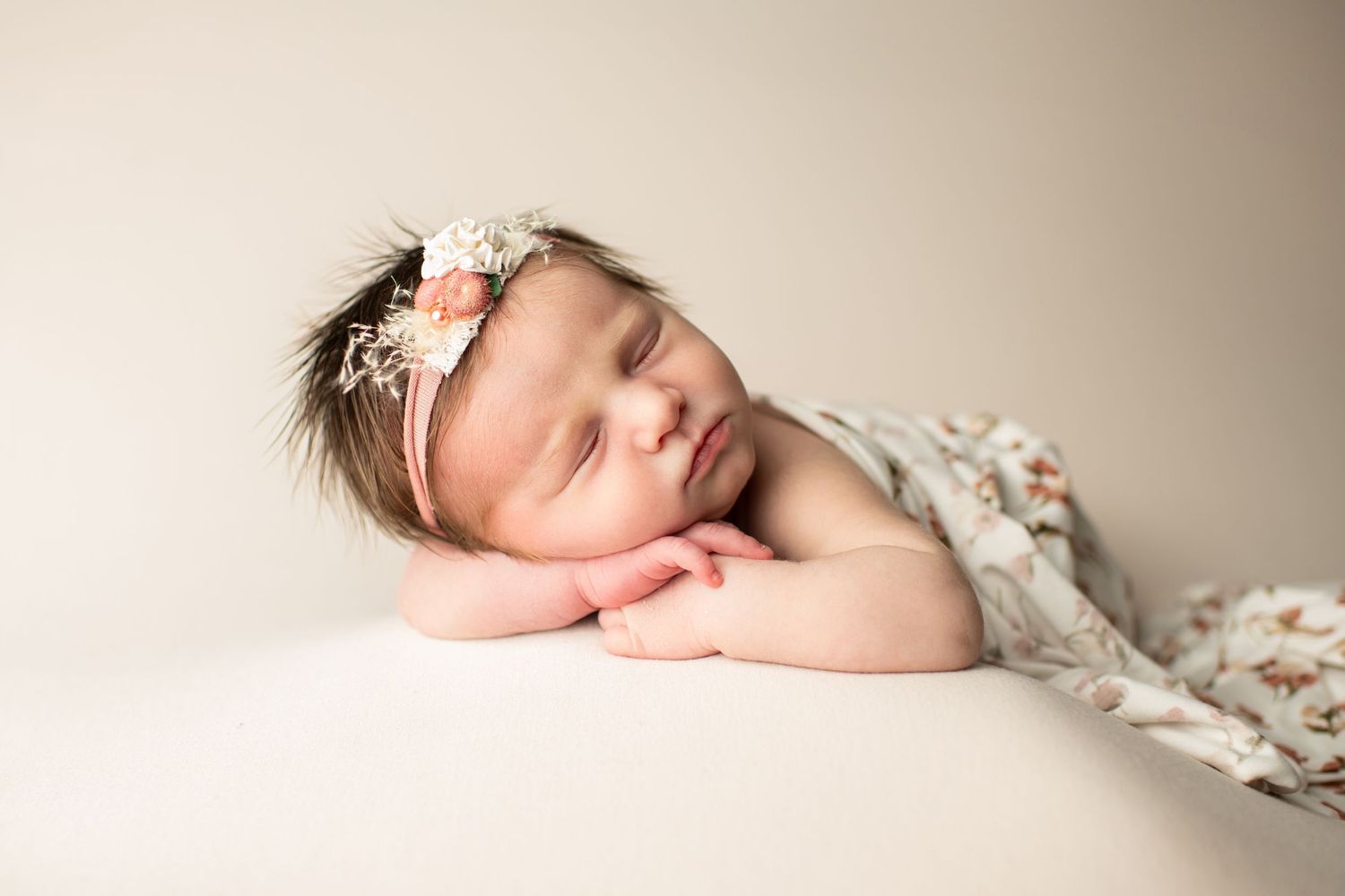 A peaceful newborn baby sleeps on a cream surface wearing a floral headband and wrapped in a delicate floral fabric.