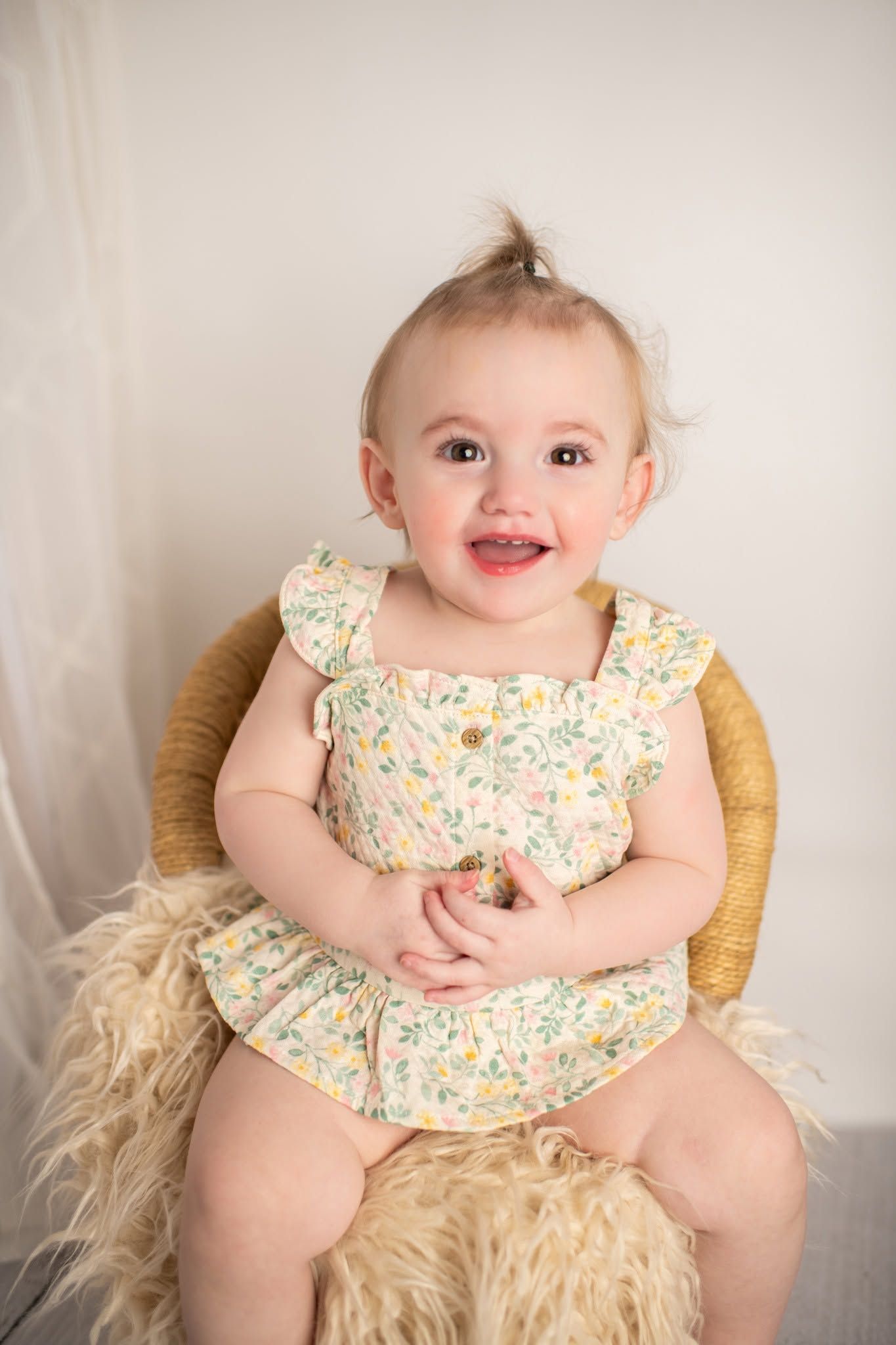 Smiling baby girl in floral romper sitting on wicker chair with fluffy rug, studio portrait photo.