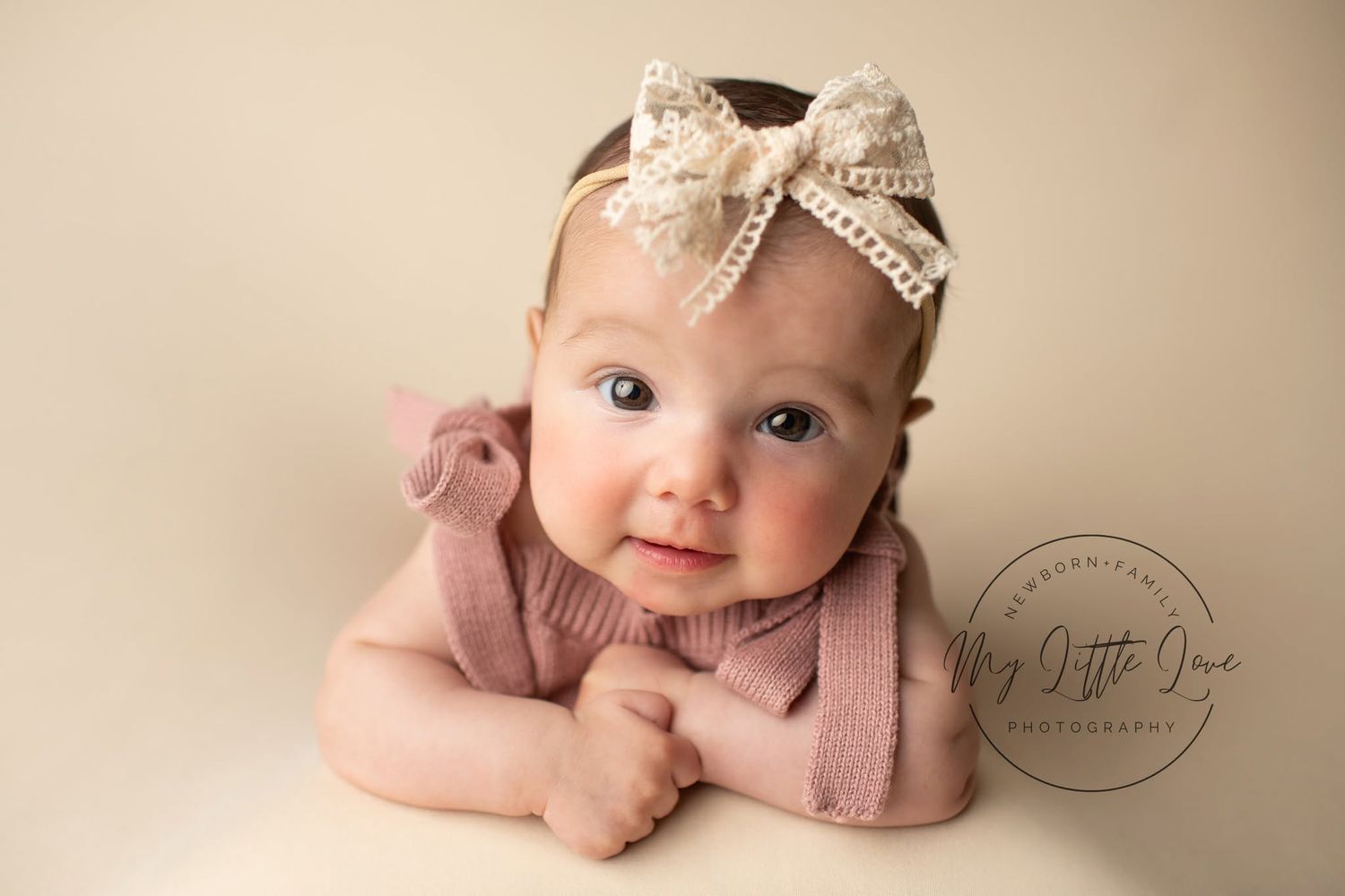 Adorable baby girl in pink romper and lace bow headband poses for a professional newborn photography portrait.