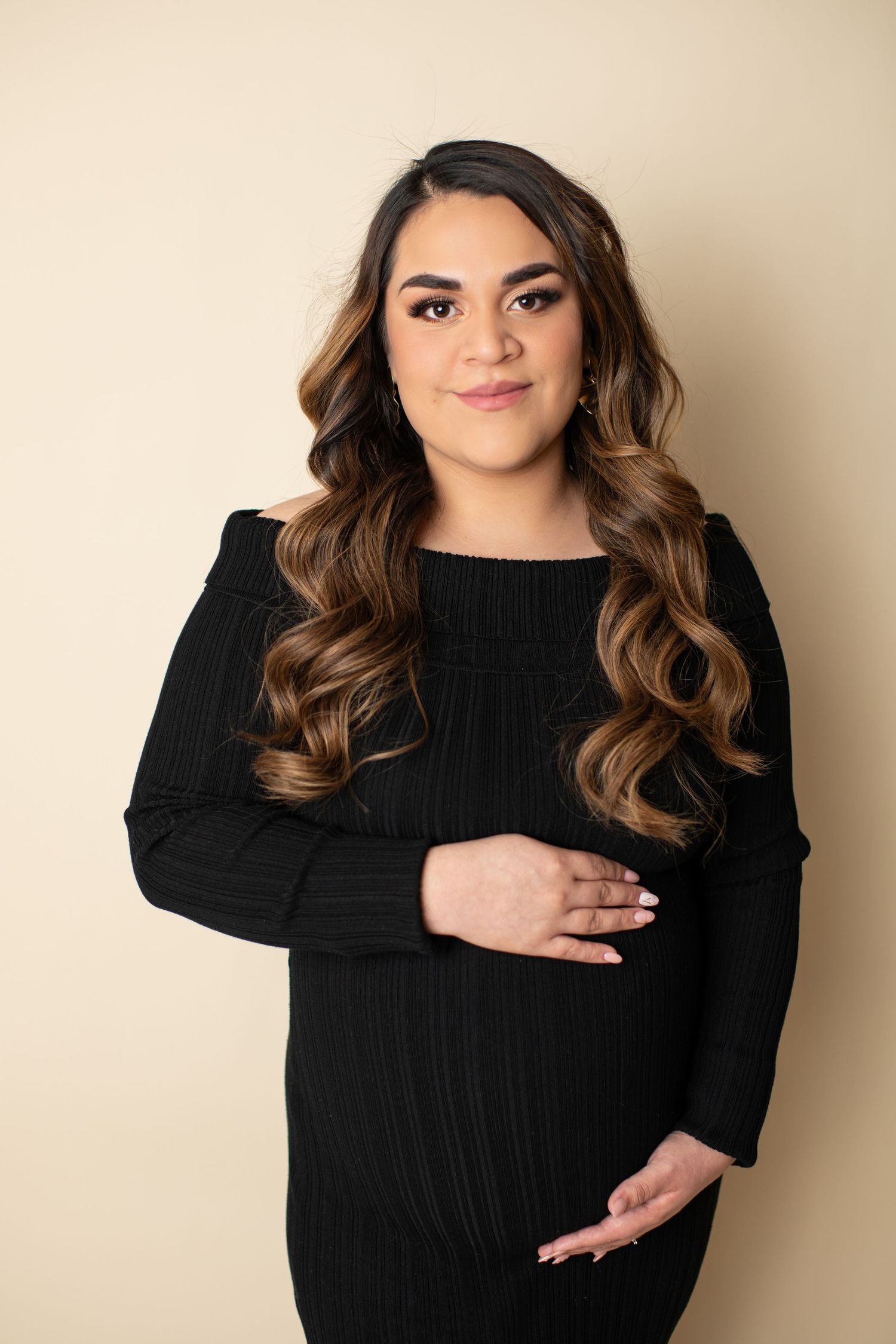 Portrait of someone wearing a black sweater against beige background with long wavy brown hair and natural makeup.