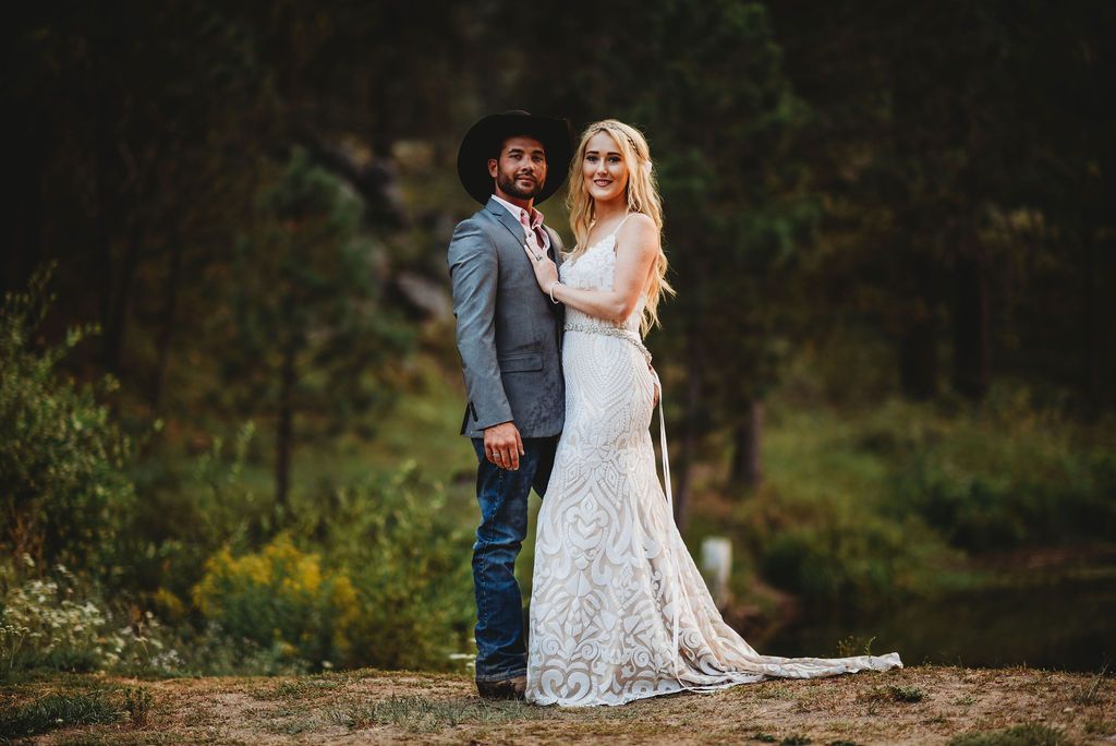 A couple stands together outdoors on a dirt path surrounded by greenery during a wedding photoshoot.