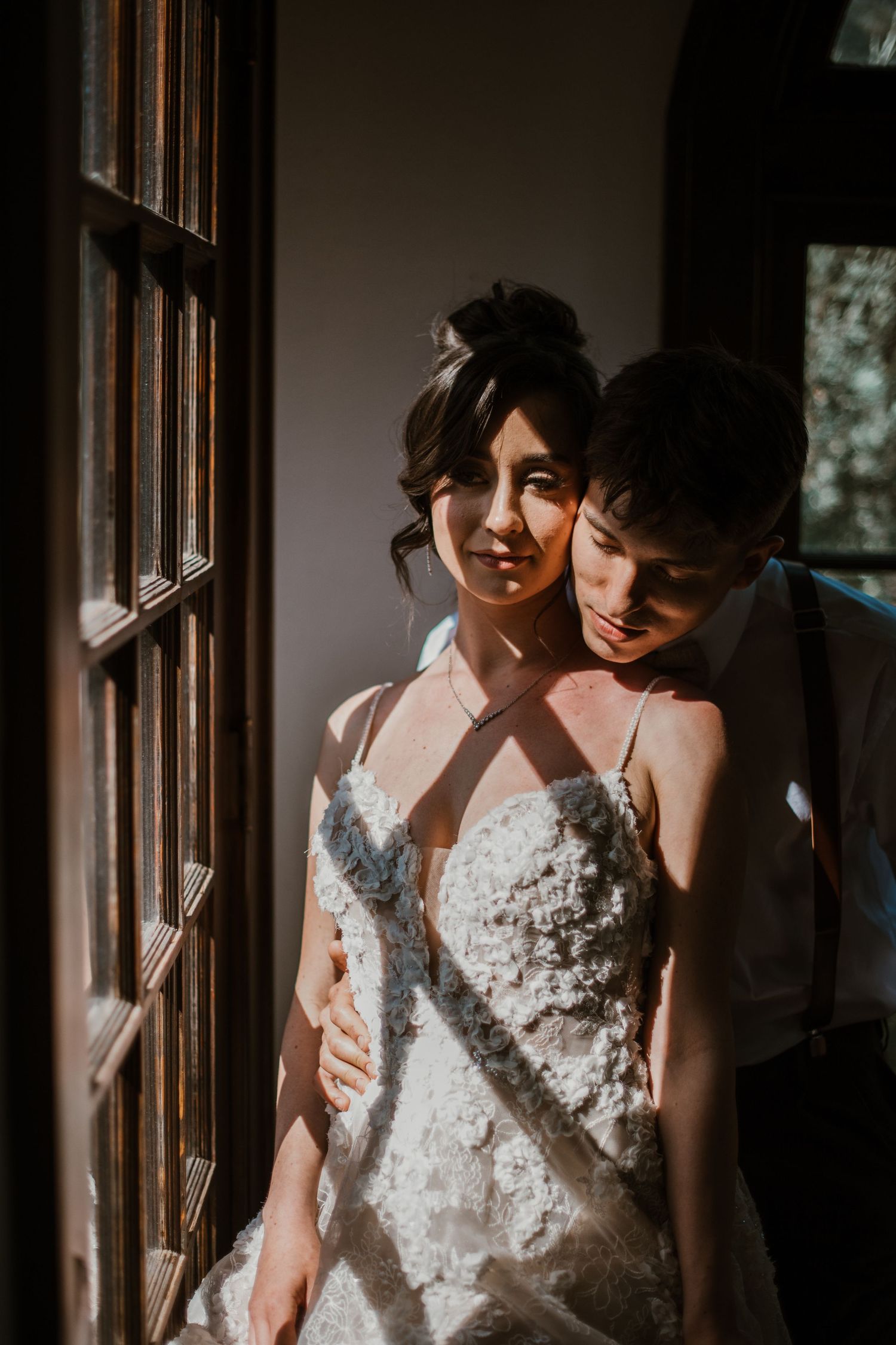 Couple in intimate lighting by window with sunlit white lace wedding dress creating dramatic shadows and mood.