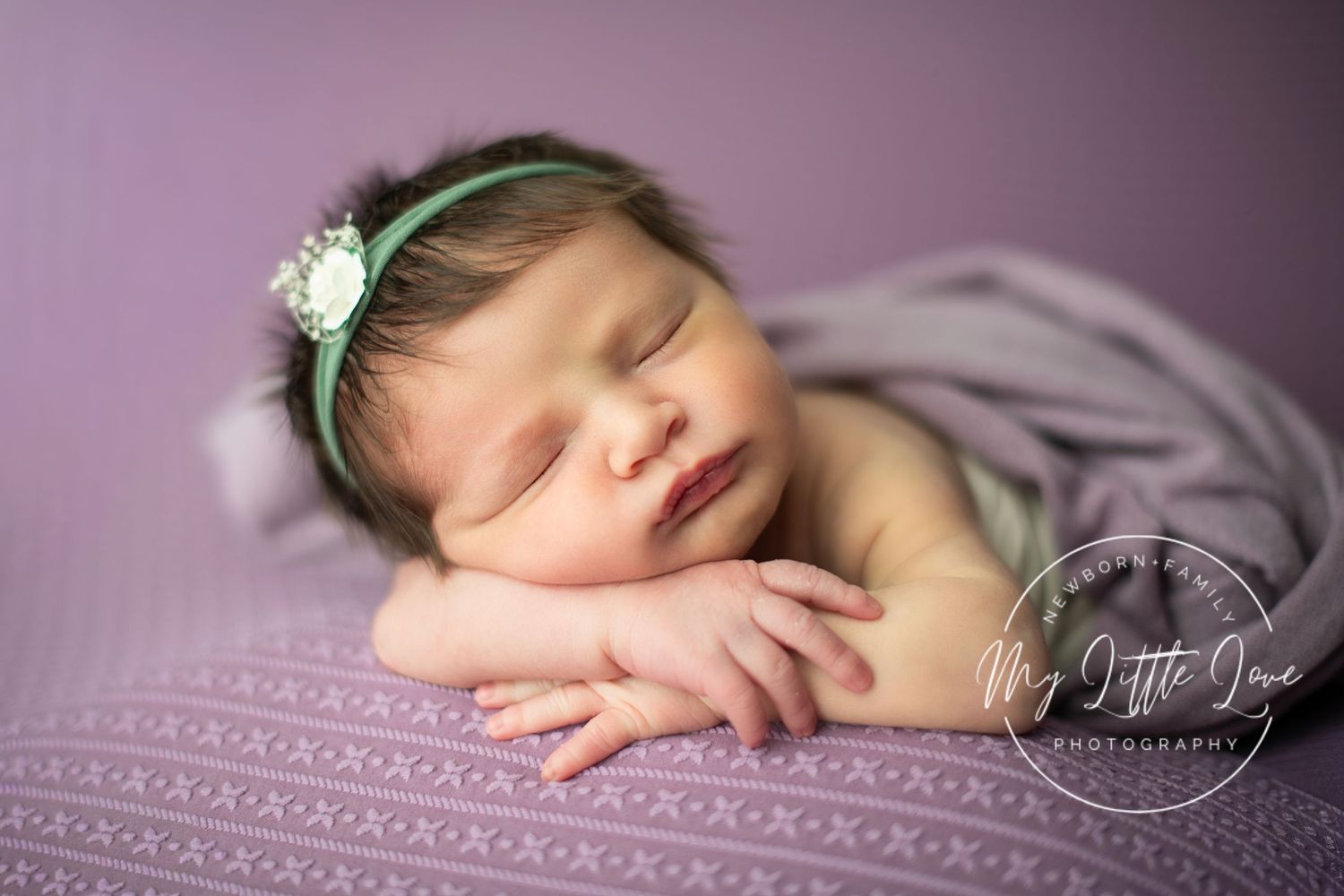 Sleeping newborn wearing delicate floral headband rests peacefully on soft purple textured blanket in professional portrait.