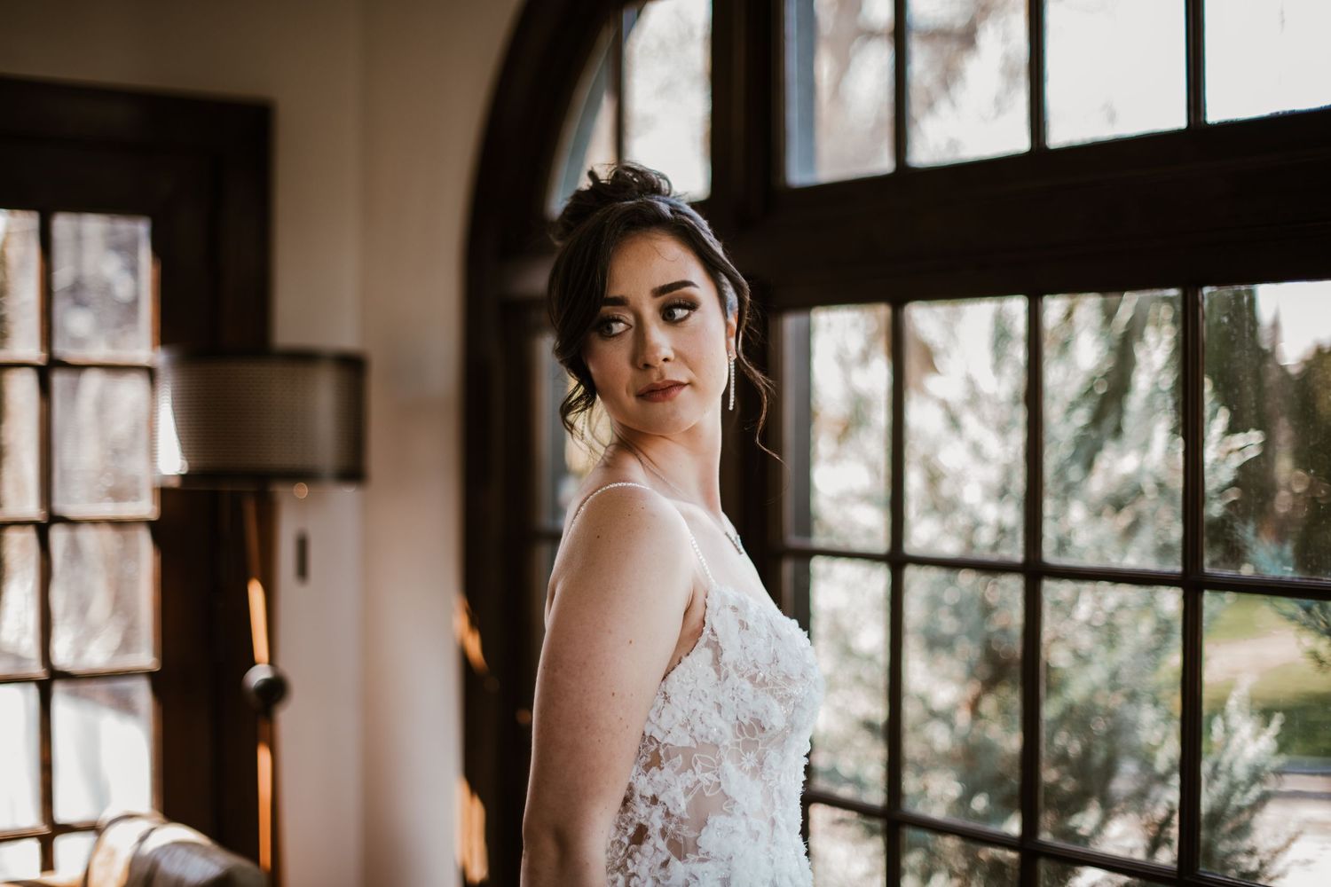 Person wearing white lace wedding dress poses near large arched windows in elegant indoor setting.