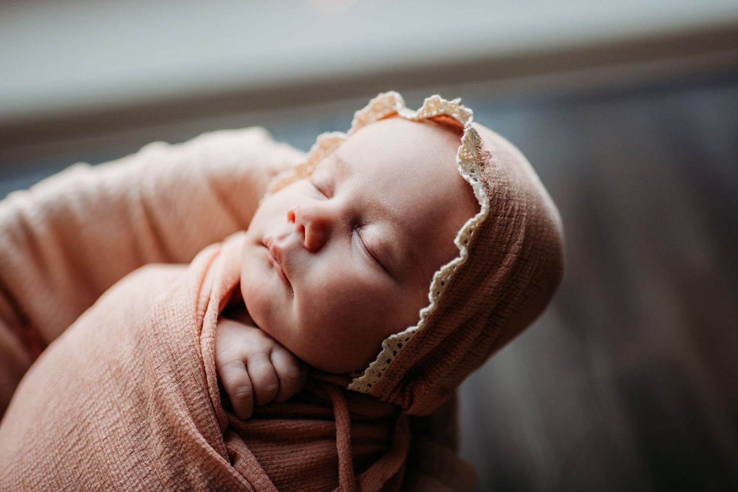 Sleeping newborn in soft brown bonnet and swaddle wrap lying peacefully during portrait session.