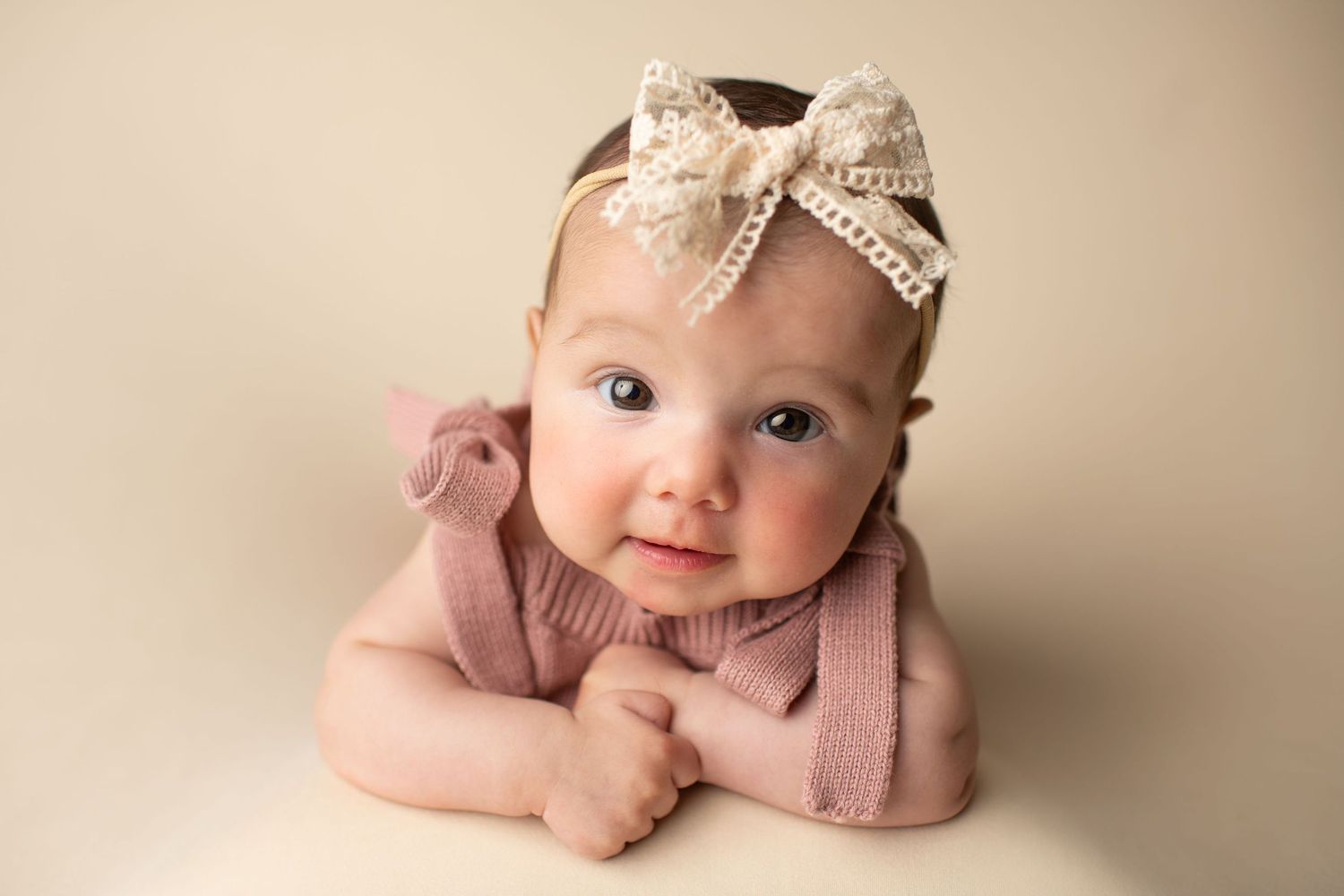 Baby wearing pink outfit and lace bow headband lying on beige backdrop during portrait session.