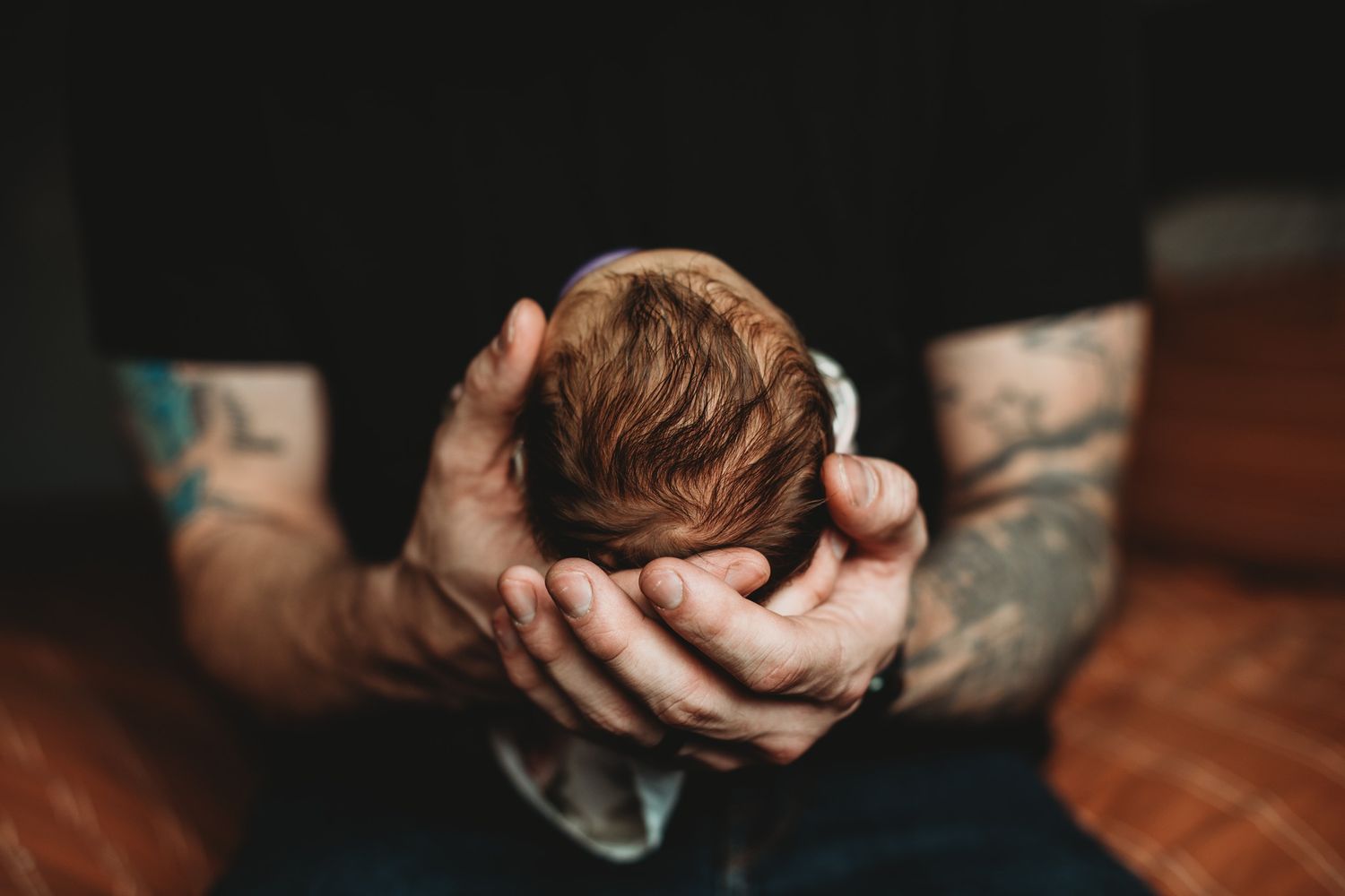 Tattooed hands gently cradling a newborn baby's head against a dark wooden background.