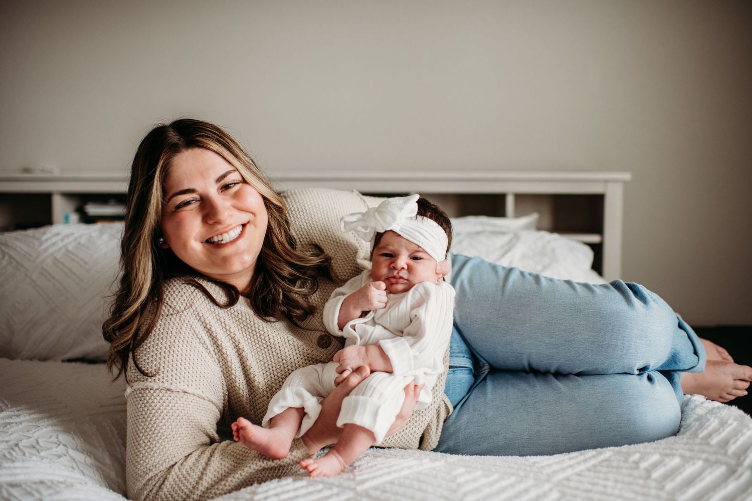A mother in a beige sweater lies on a bed with a newborn baby, both smiling in a cozy bedroom setting.