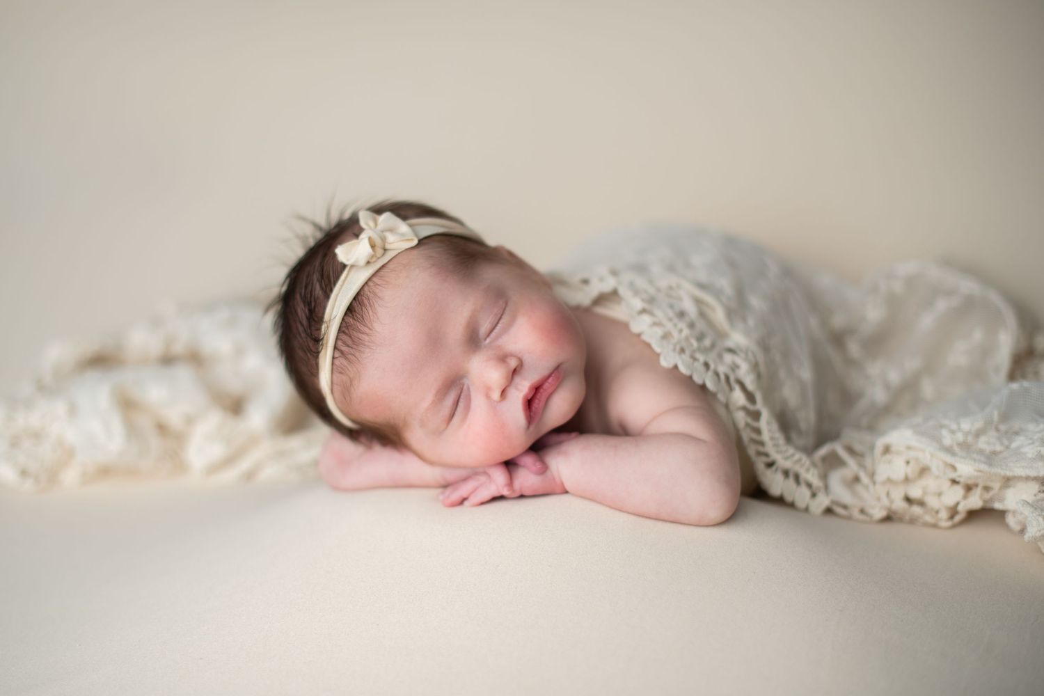 Sleeping newborn baby girl wearing a delicate lace headband, wrapped in cream lace fabric on a soft white background.
