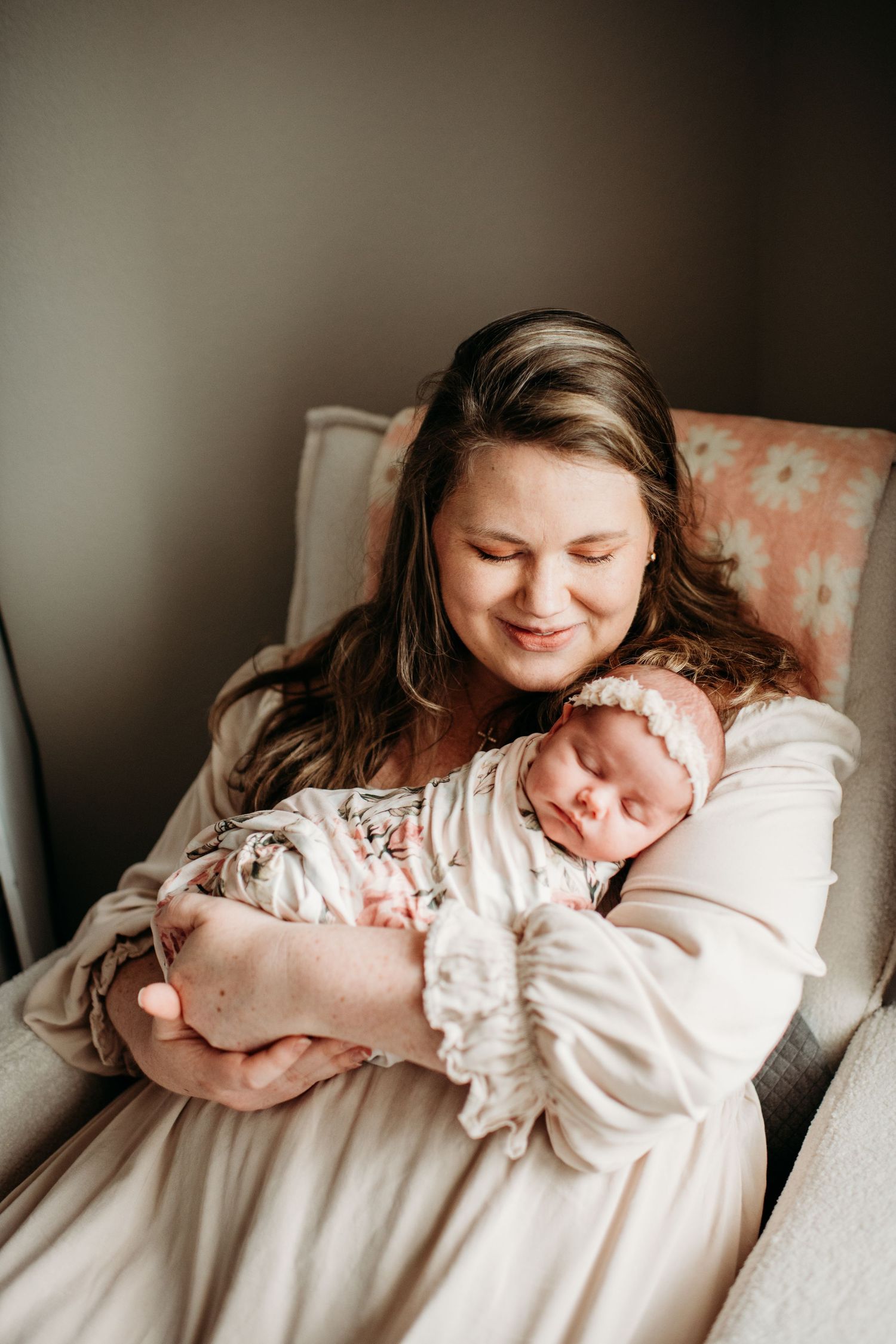 A tender moment shows mother and newborn baby snuggling in a cream-colored armchair with soft lighting.