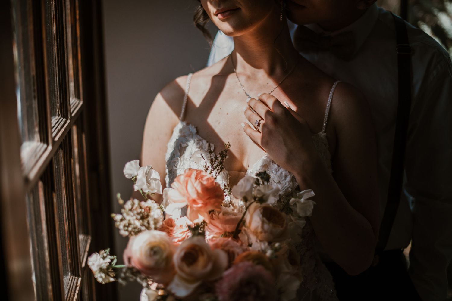 A romantic wedding bouquet of soft peach and white flowers is held near a window in dreamy natural light.