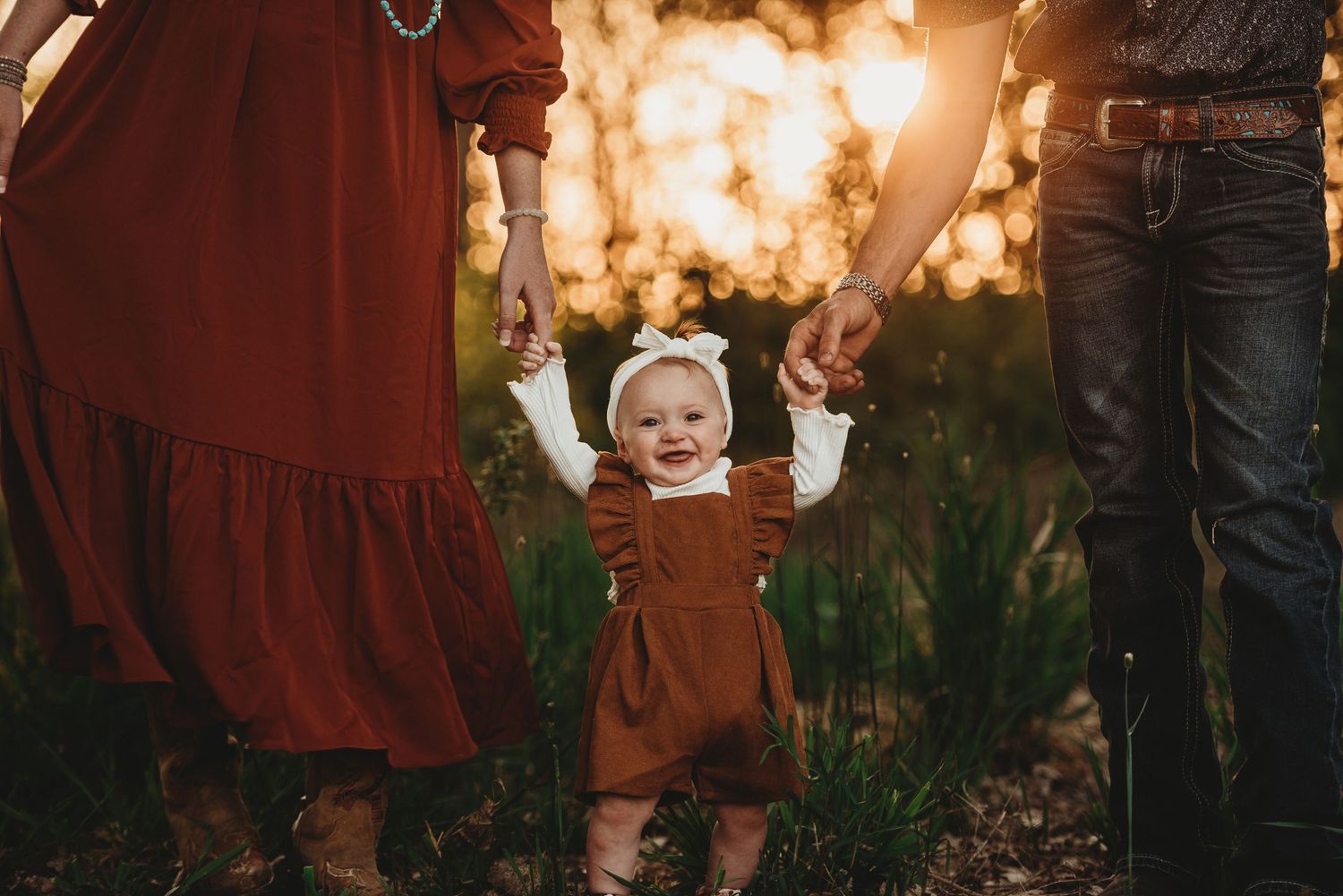 Small child in brown overalls standing between parents against golden sunset backdrop.