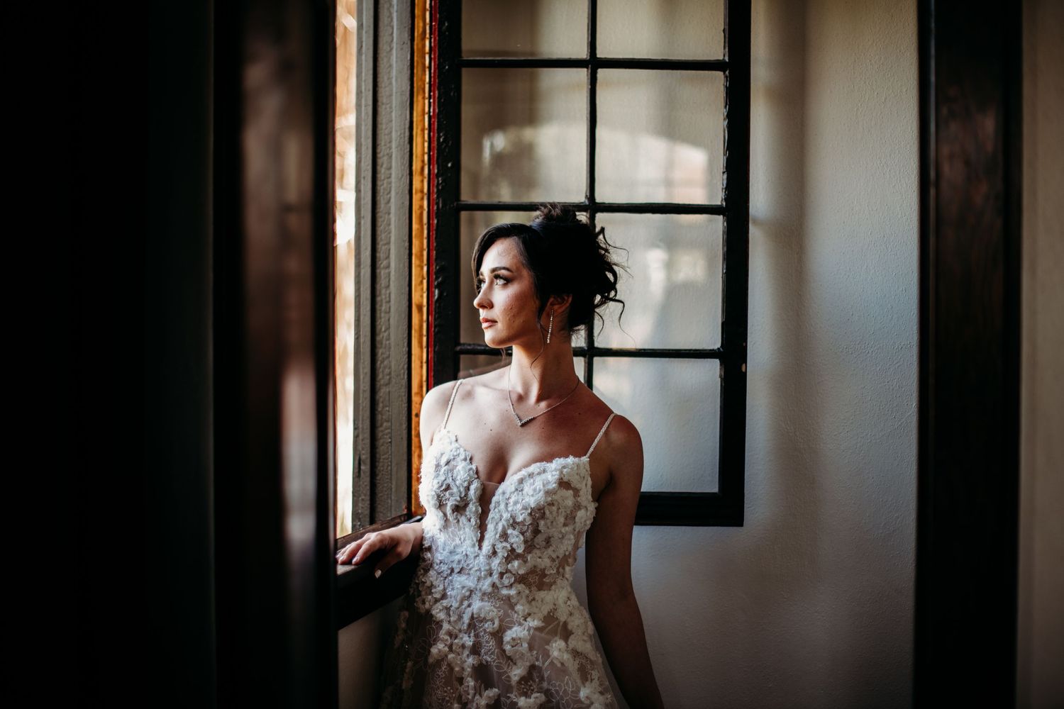 A bride in a delicate floral lace wedding gown gazes thoughtfully out of a window in soft natural light.