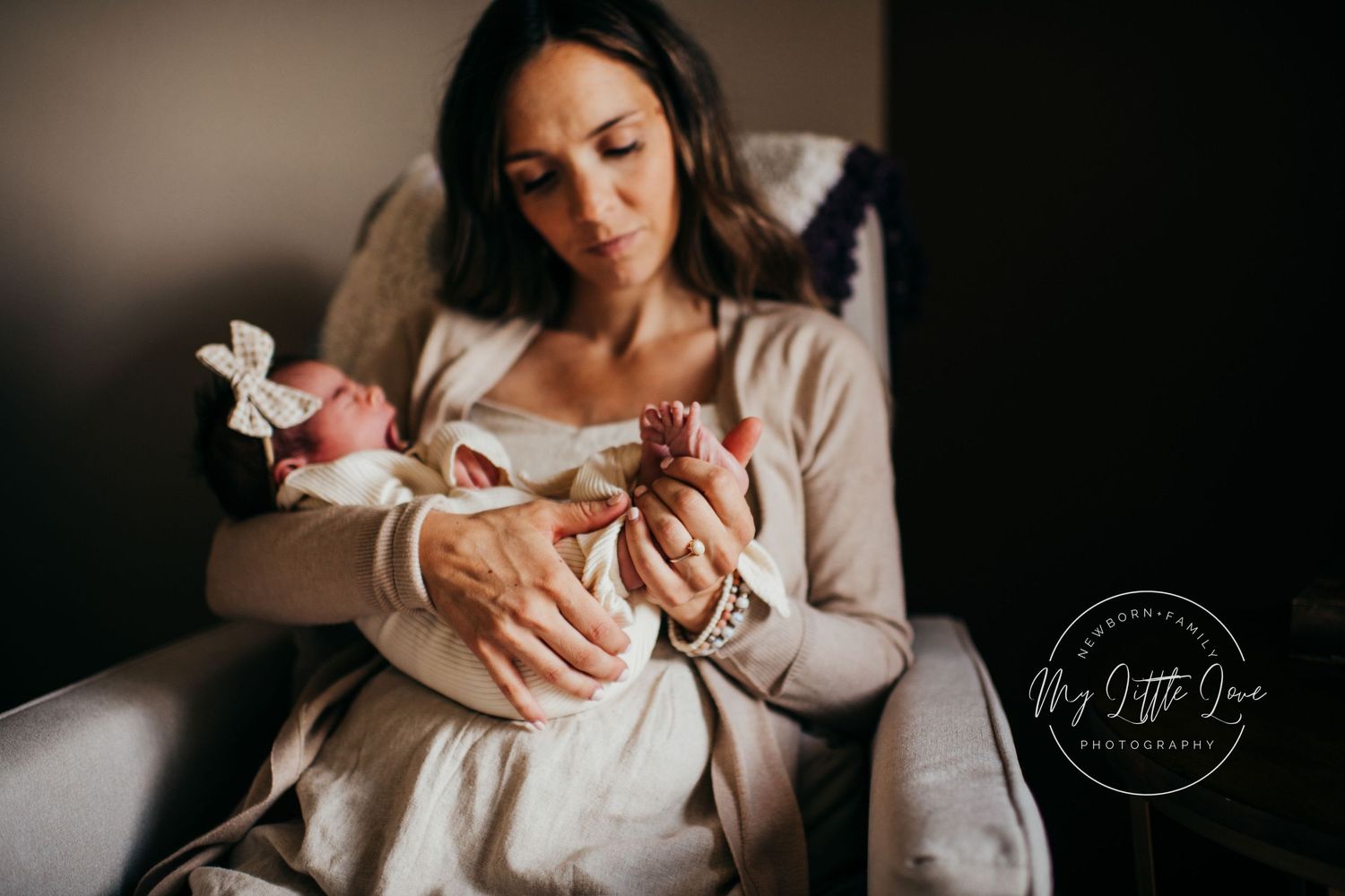 Mother in neutral sweater holding newborn baby in cozy armchair setting.