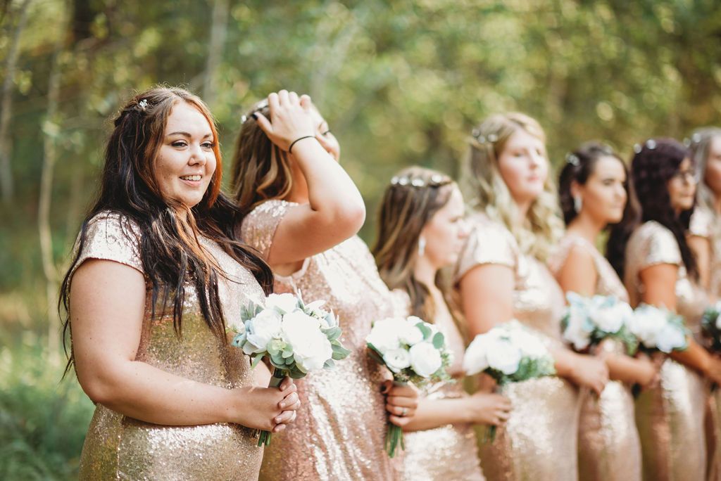 Bridesmaids in sparkly gold dresses laughing together while holding white bouquets during outdoor wedding photography.