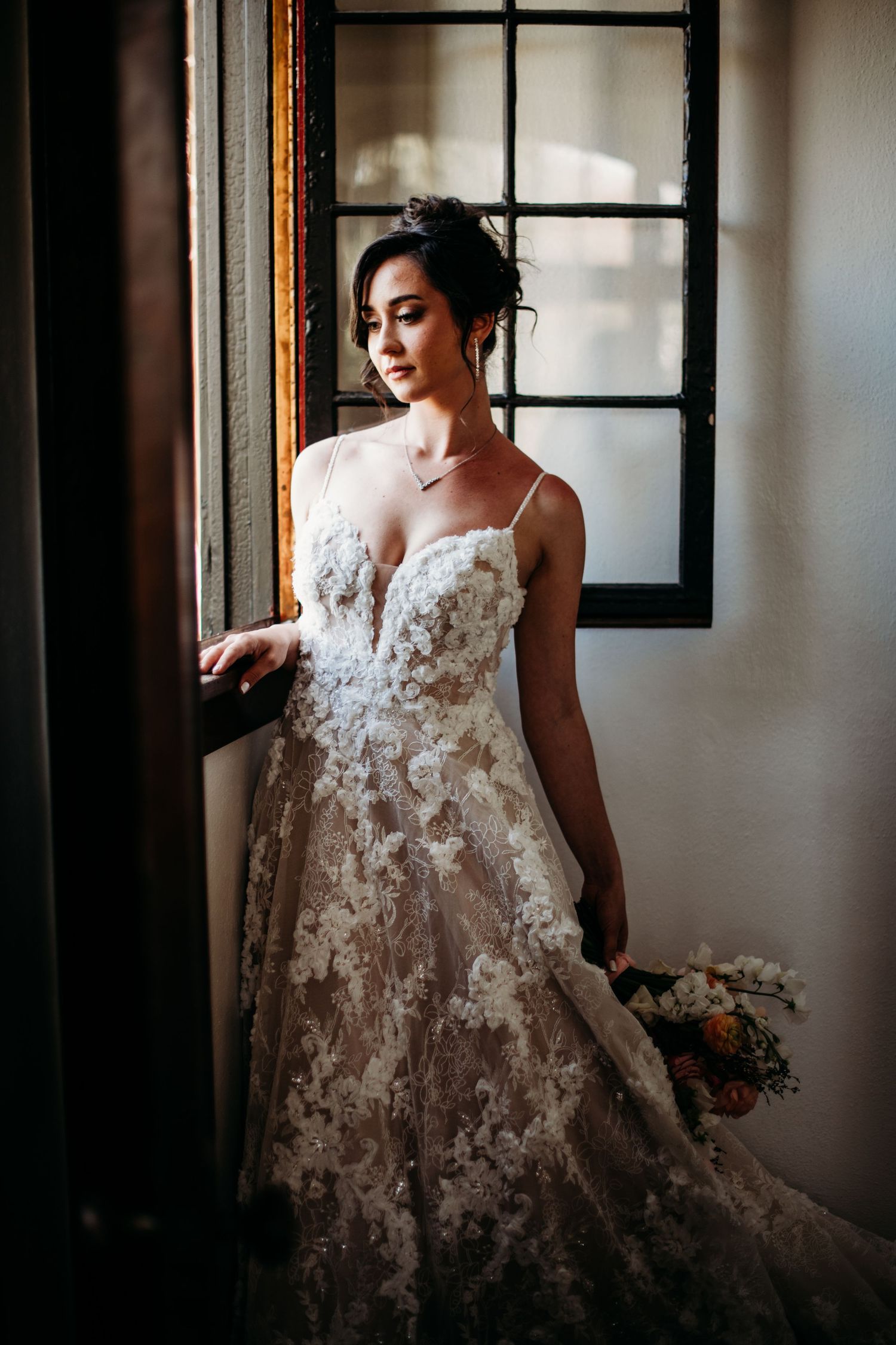 Elegant lace wedding gown with floral appliques photographed by window in romantic natural lighting.