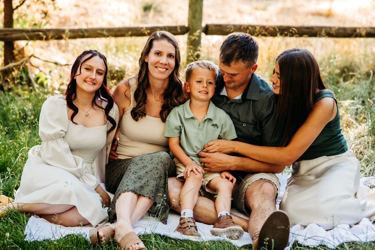 A happy family of five smiles together outdoors on a blanket, surrounded by lush greenery and a wooden fence.