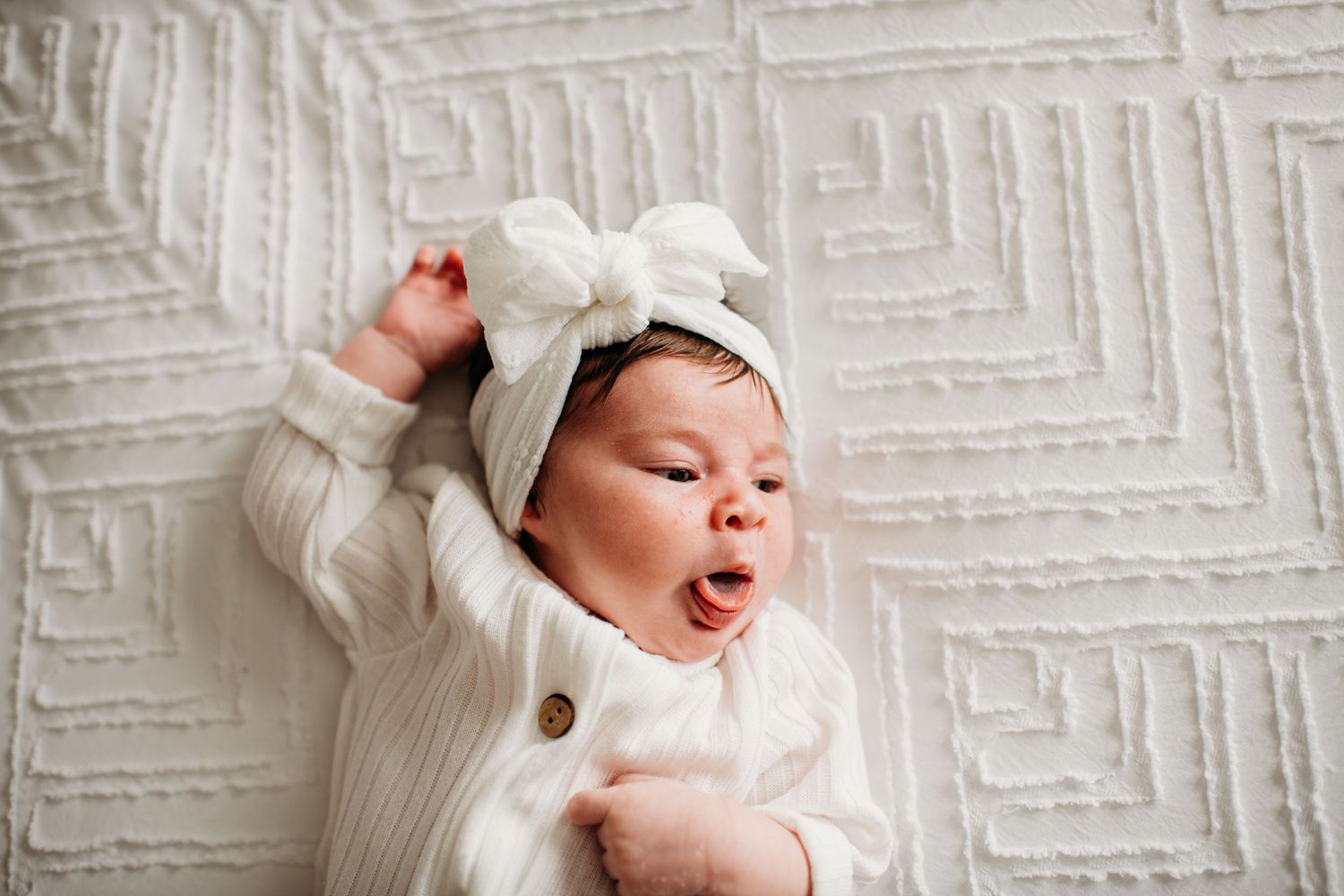 Baby wearing white sweater and hat lies on textured white blanket while yawning.