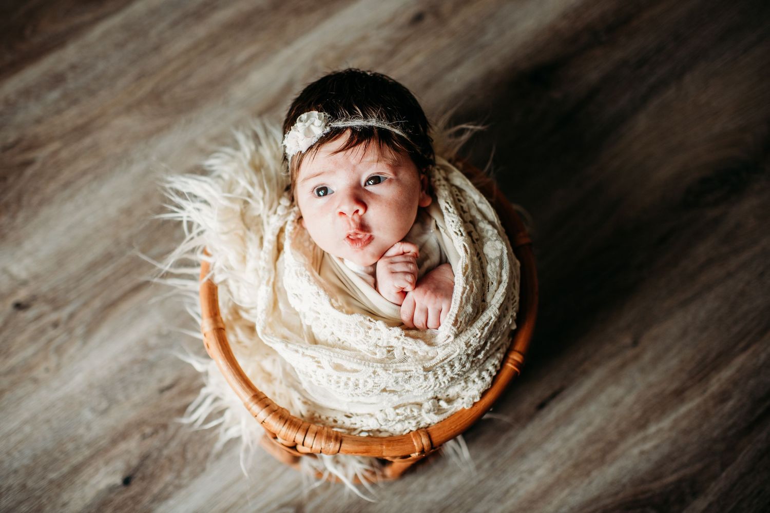 A newborn baby swaddled in soft cream fabric nestles in a round wooden bowl on a gray floor.