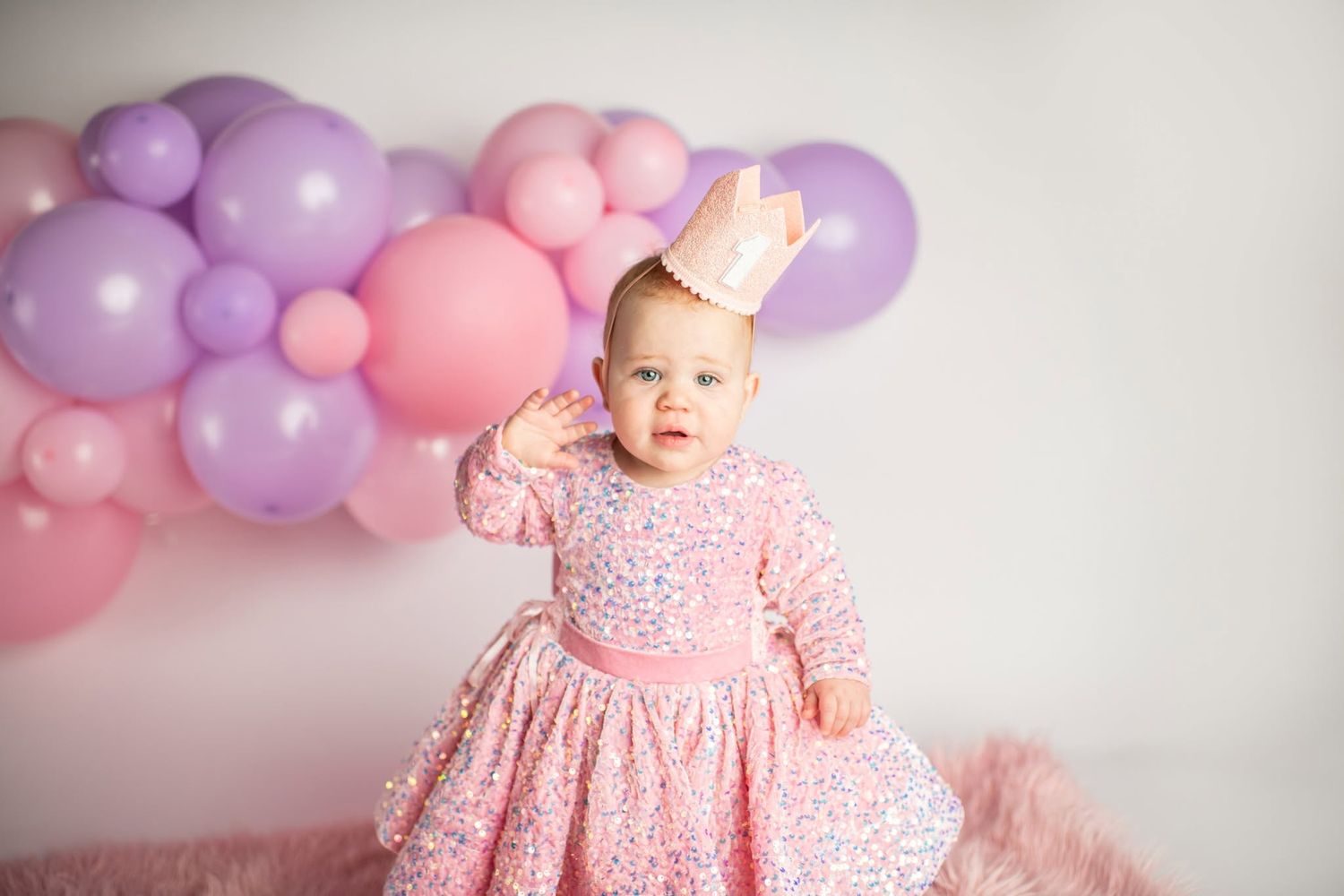 A baby girl in a pink sequin dress and crown poses in front of colorful pink and purple balloons at her first birthday.