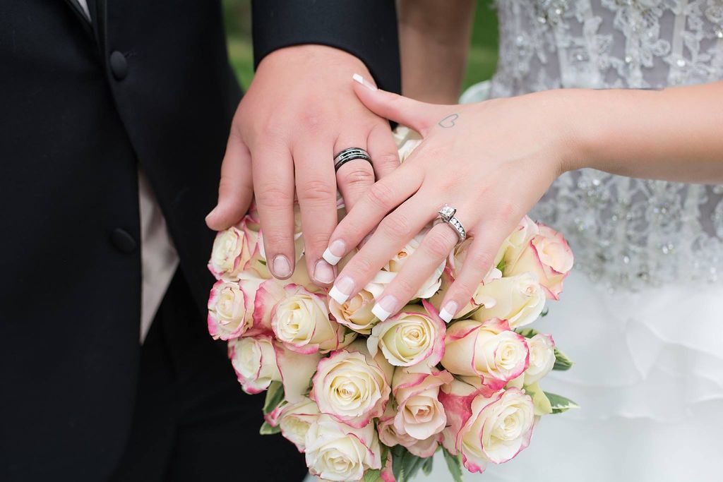Close-up shot of hands with wedding rings resting on a bouquet of blush pink roses.