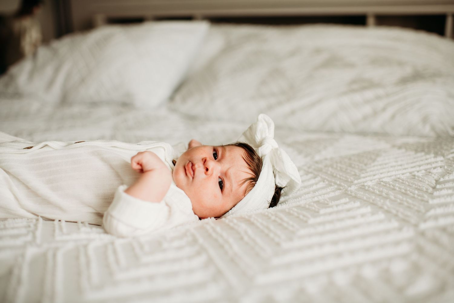 A happy baby in white clothing lies on textured bedding while looking up with a bright smile.