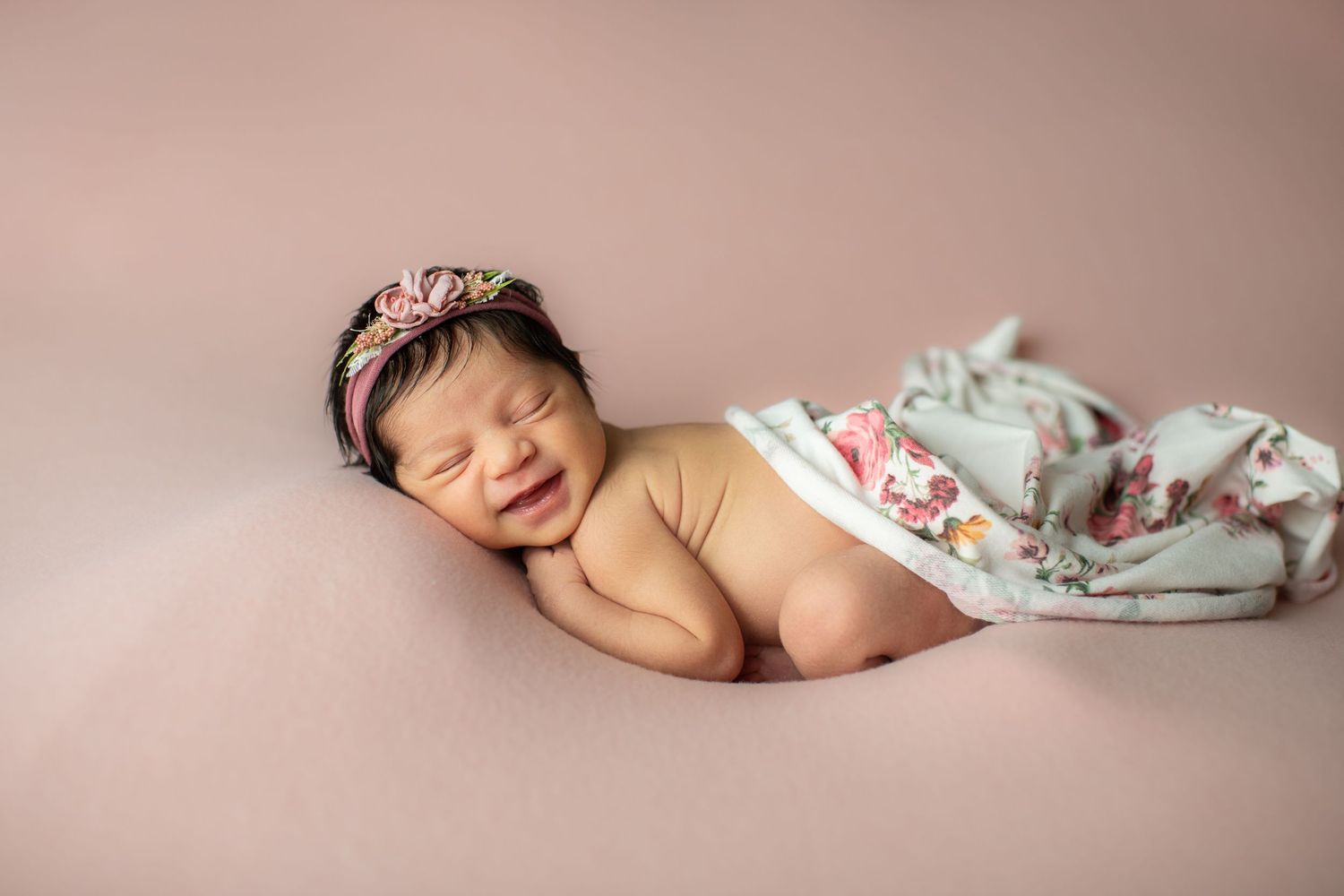 Sleeping newborn wearing a tiara headband lies on pink backdrop with floral patterned blanket.