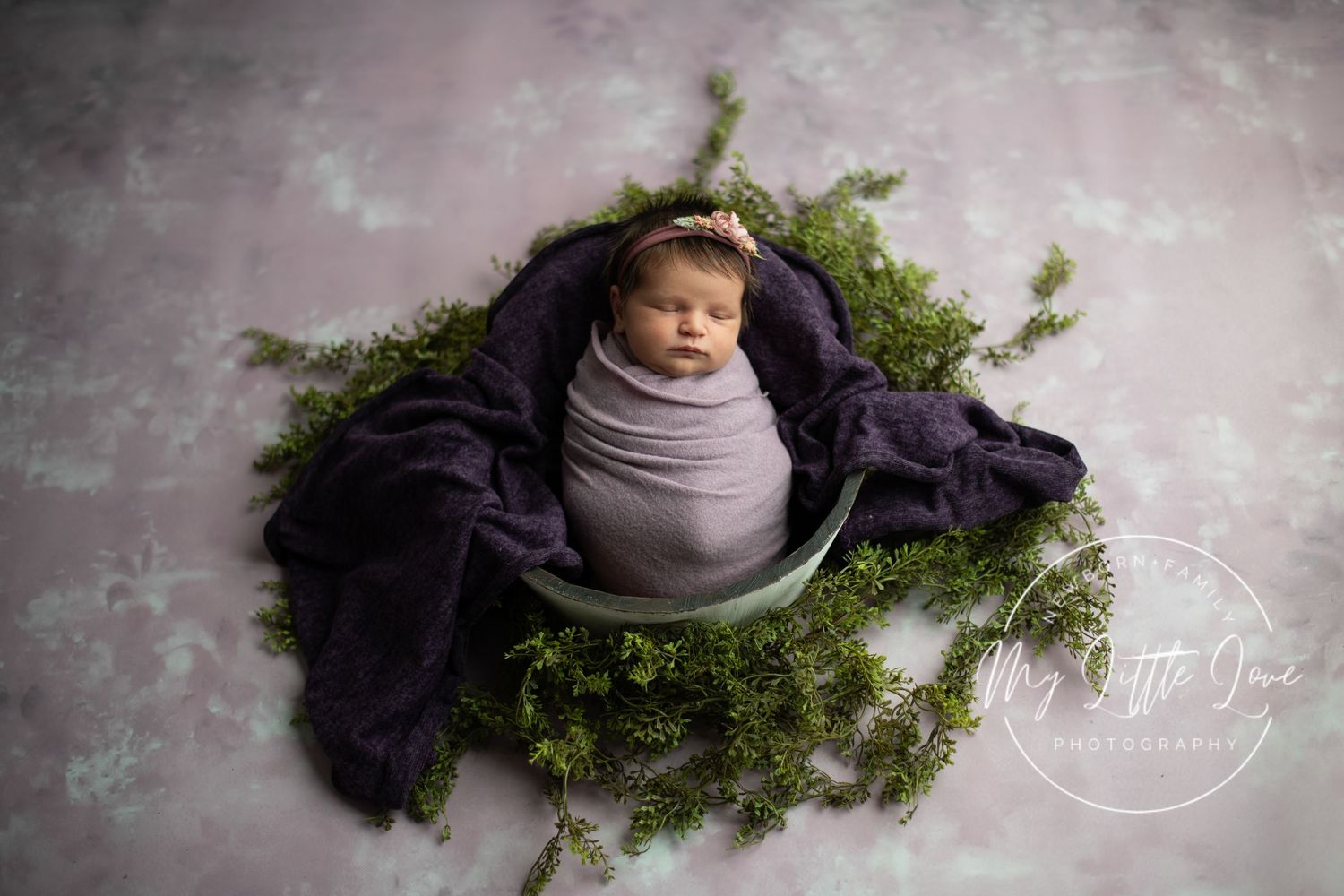 Artistic newborn photography series showing baby wrapped in purple fabric with natural greenery accents.