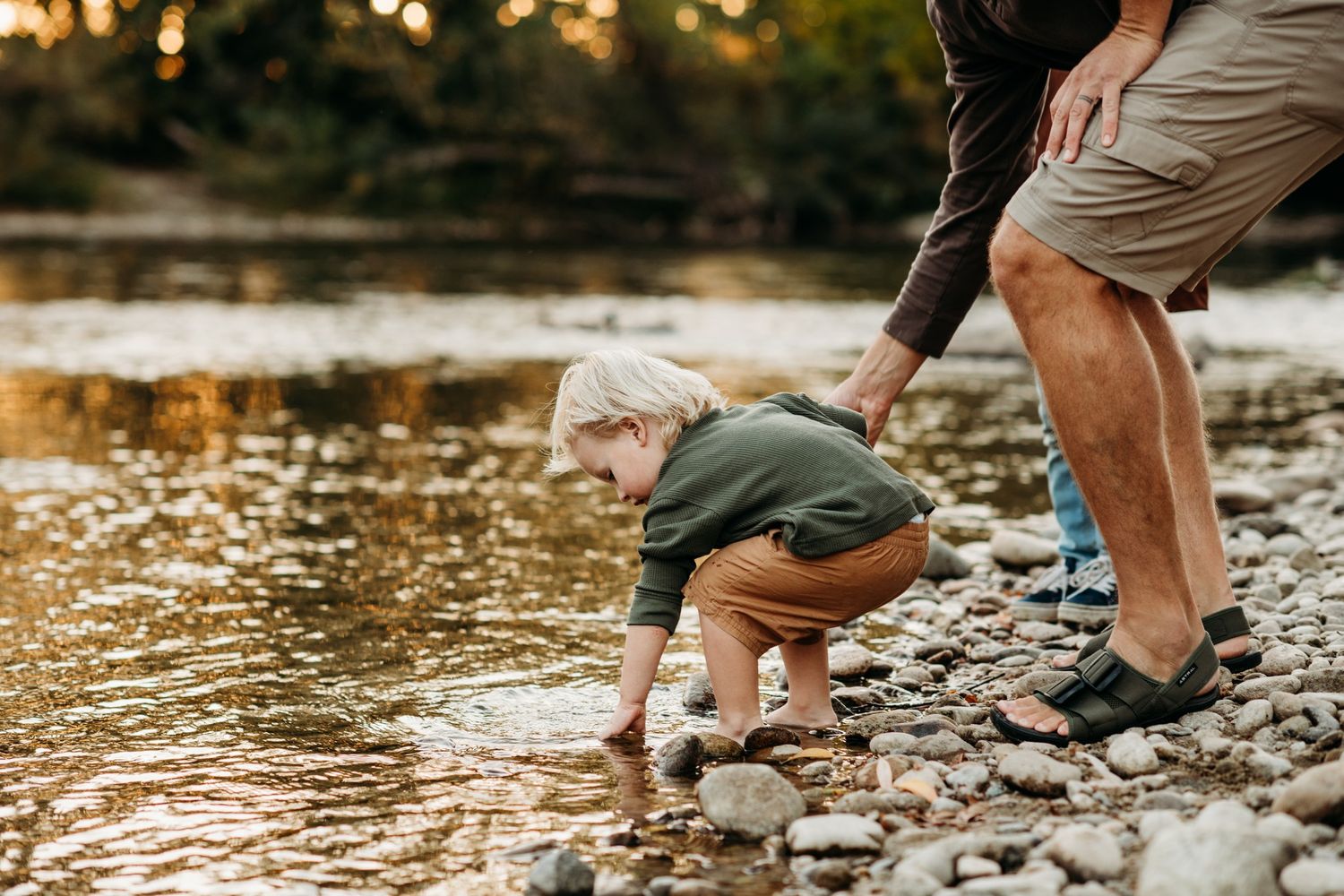 Parent and child explore rocky riverbank at sunset while collecting stones together.