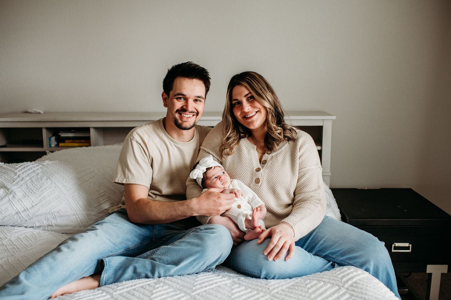 A couple sits on a bed holding their newborn baby while wearing casual neutral-colored clothing in a bright room.