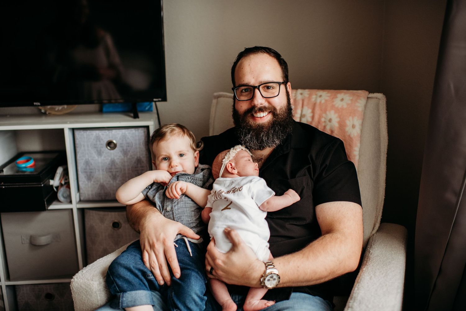 A bearded man in a black t-shirt sits in an armchair with two young children at home.