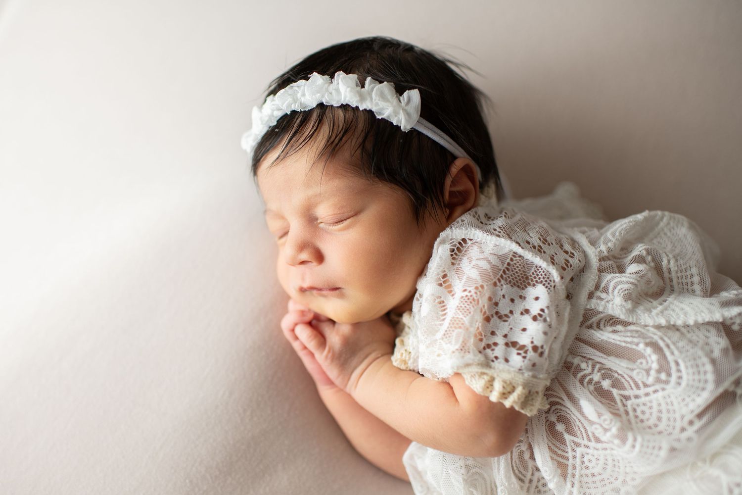 Peaceful newborn in white lace dress and dainty headband sleeps with hand tucked under chin.