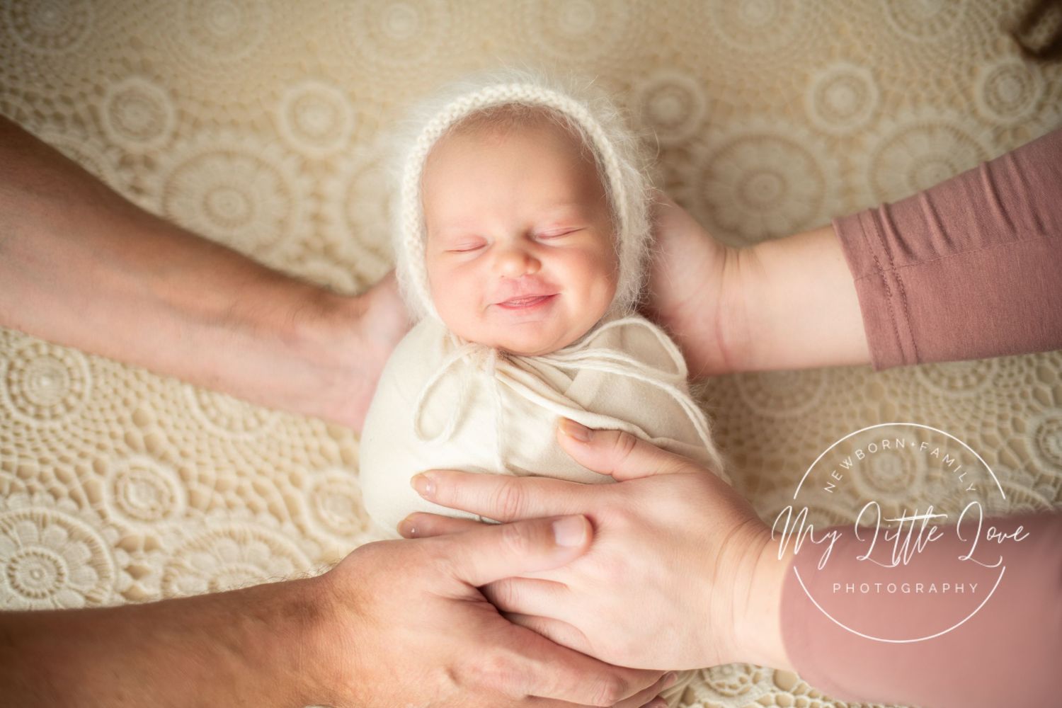 A sleeping newborn baby swaddled in white fabric is gently cradled by adult hands against a beige textured background.