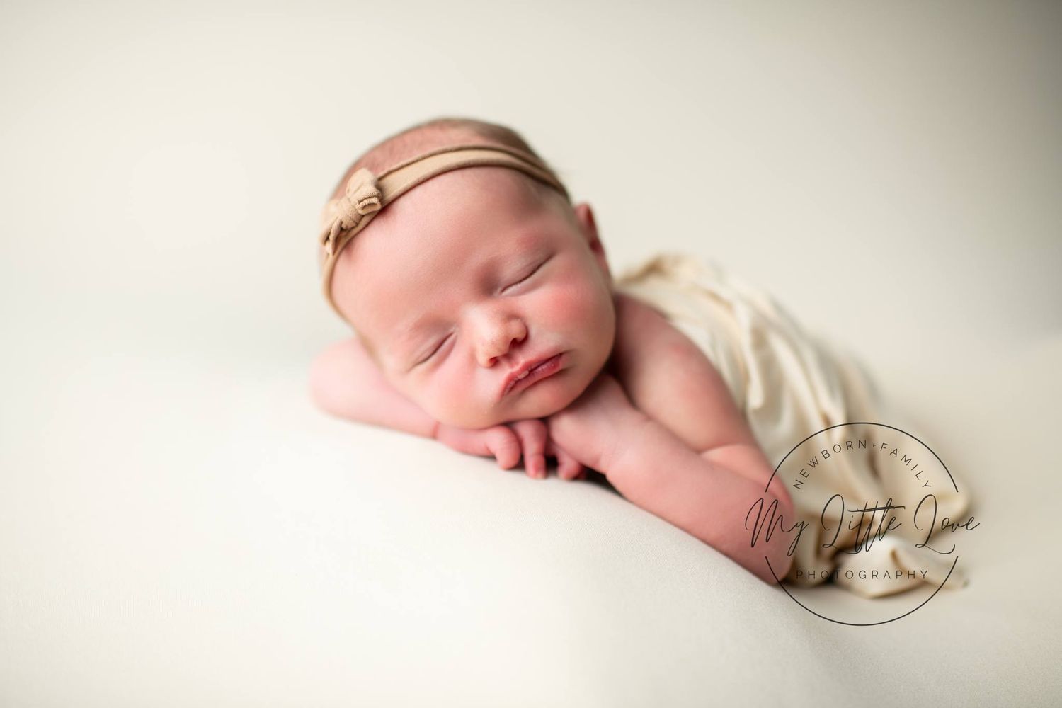 A sleeping newborn baby wearing a delicate headband on soft white fabric.