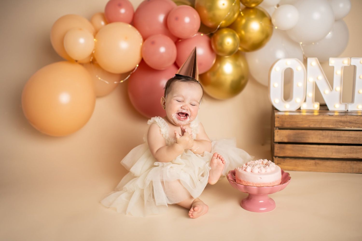 Baby girl in white dress crying at her first birthday cake smash with pink and gold balloons.