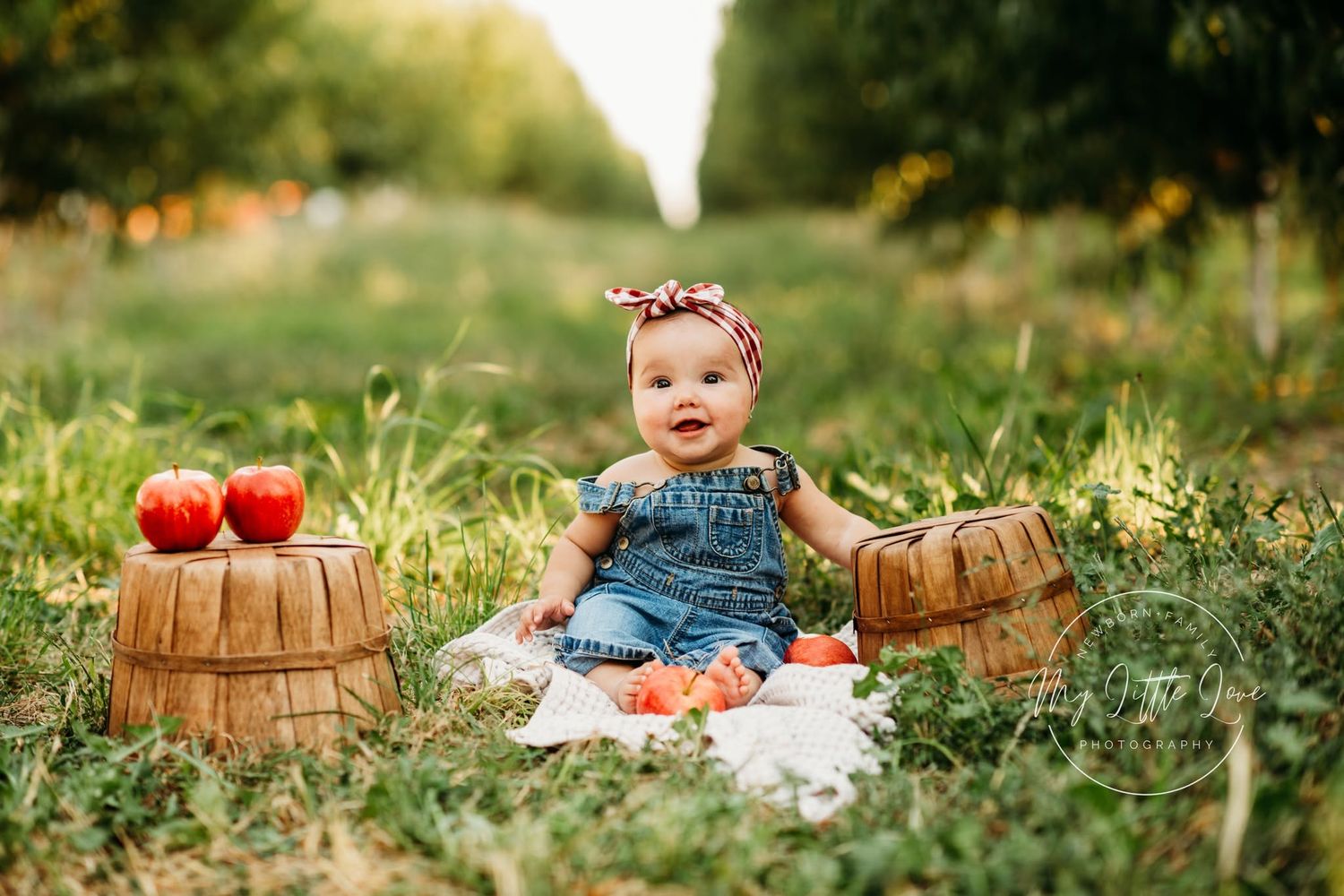 Baby girl in denim romper sits on blanket between apple baskets in an orchard for a fall photo session.