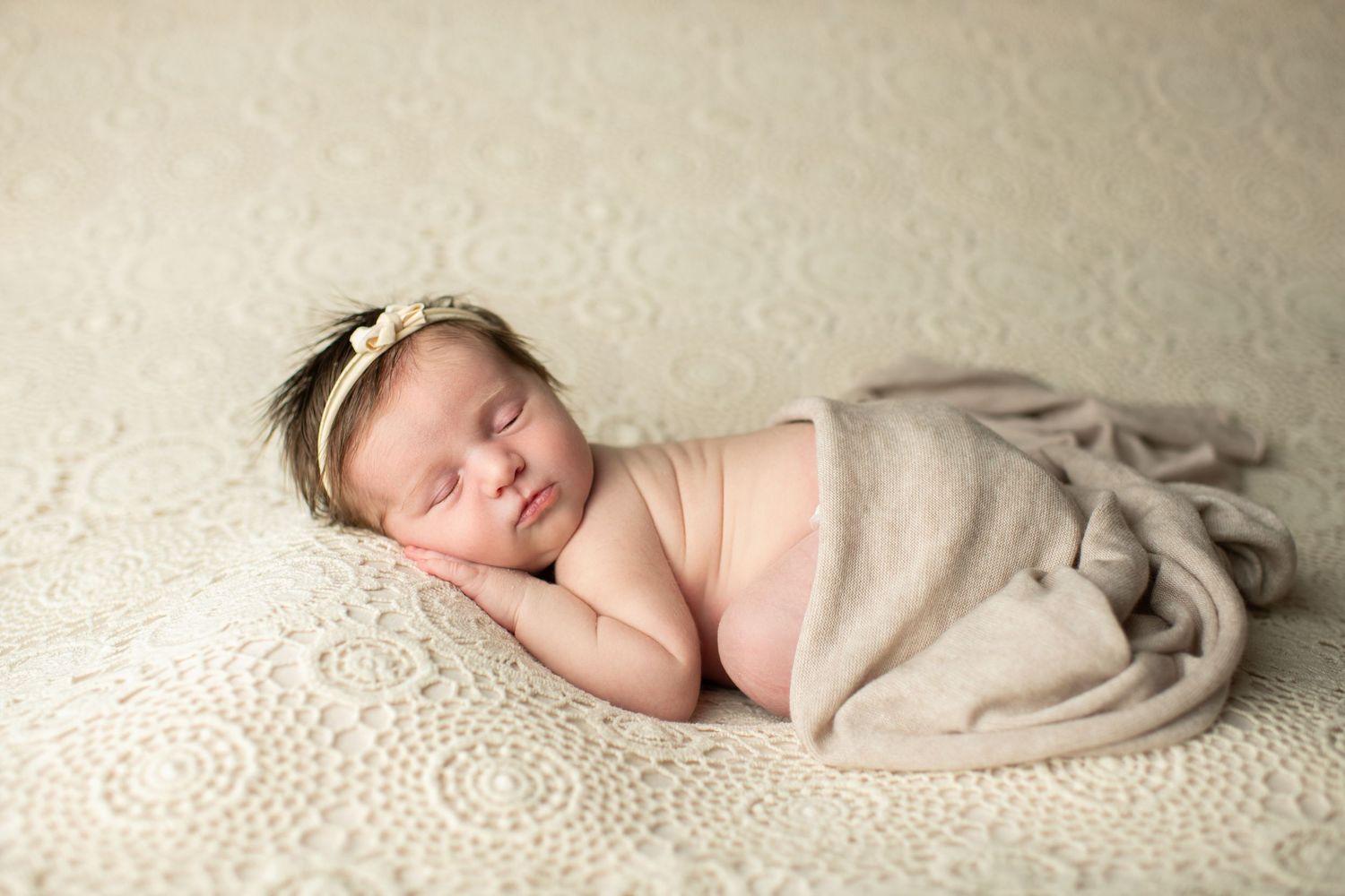 Peaceful sleeping newborn wrapped in soft beige fabric rests on textured cream lace blanket.