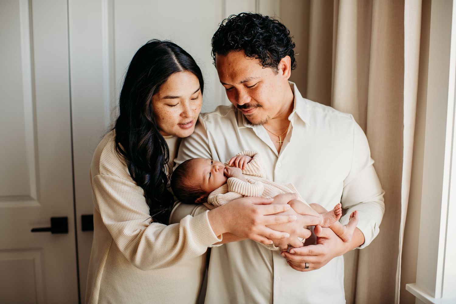 Parents in cream-colored robes cradle their newborn baby tenderly at home in a warm intimate moment together.