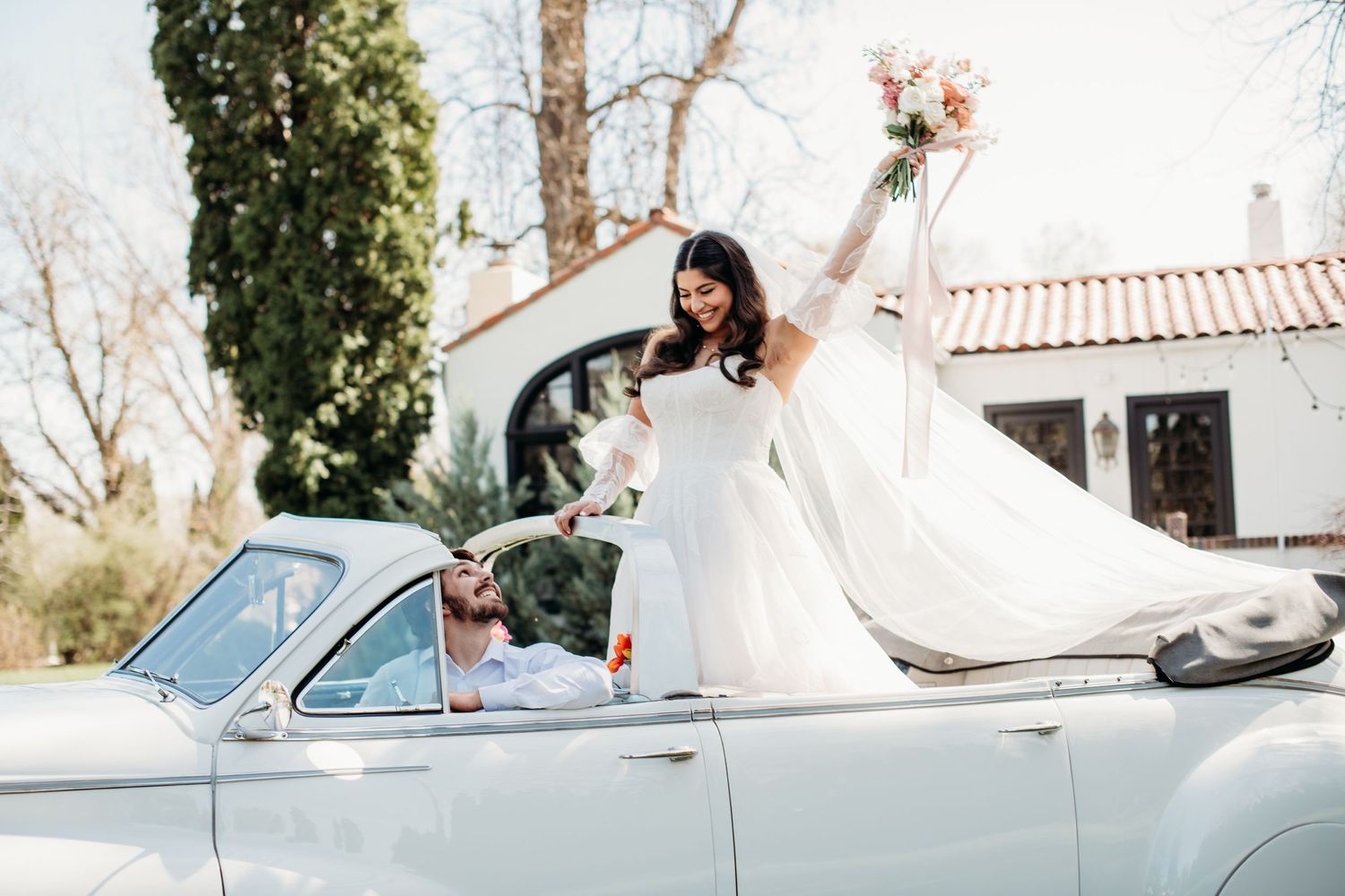 A vintage convertible car adorned with wedding decor features someone in a flowing white wedding dress raising a bouquet joyfully.