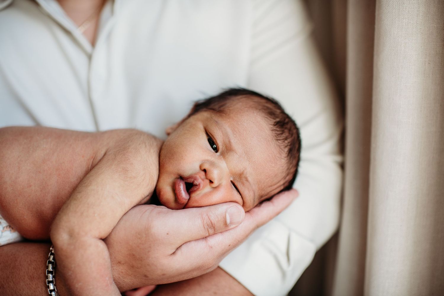 A newborn baby rests peacefully while being cradled in someone's arms next to a window.