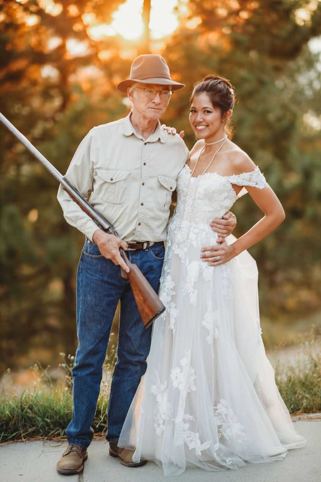 A bride in a white lace gown poses next to someone in casual attire holding a rifle during golden hour sunset.