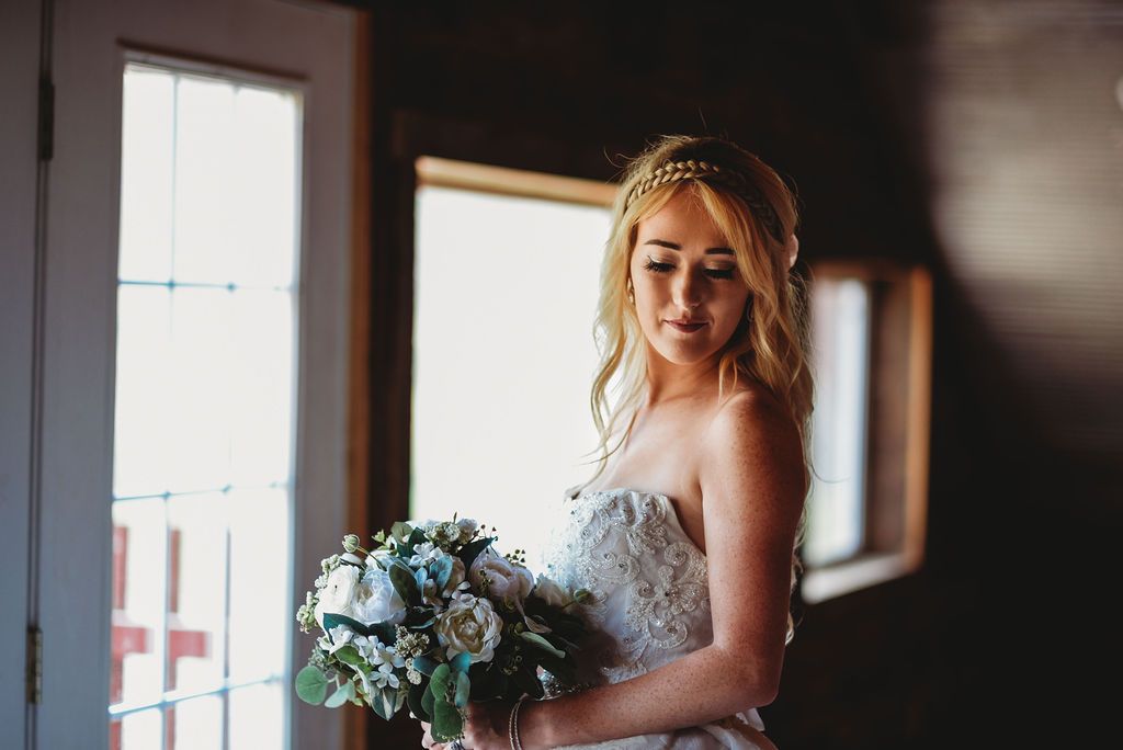 Bride in a white lace gown holding a blue and white bouquet near a sunlit window.