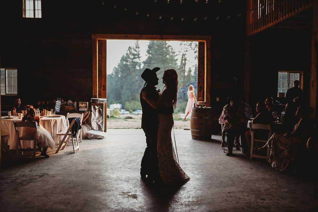 A silhouetted couple embraces in a rustic barn doorway during a wedding reception with tables and guests visible.