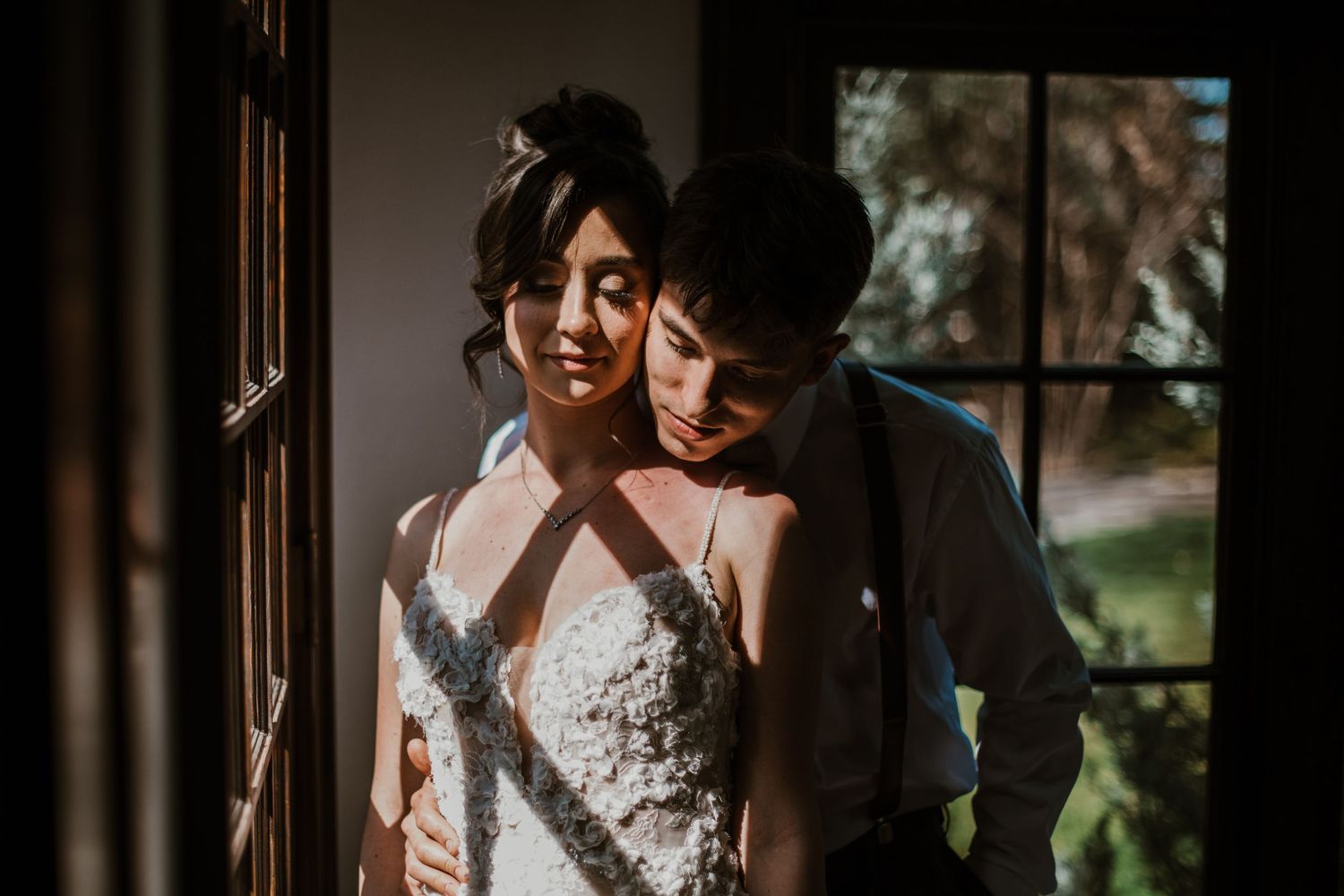 A couple shares an intimate moment by a sunlit window, the bride wearing a white halter-style lace wedding dress.